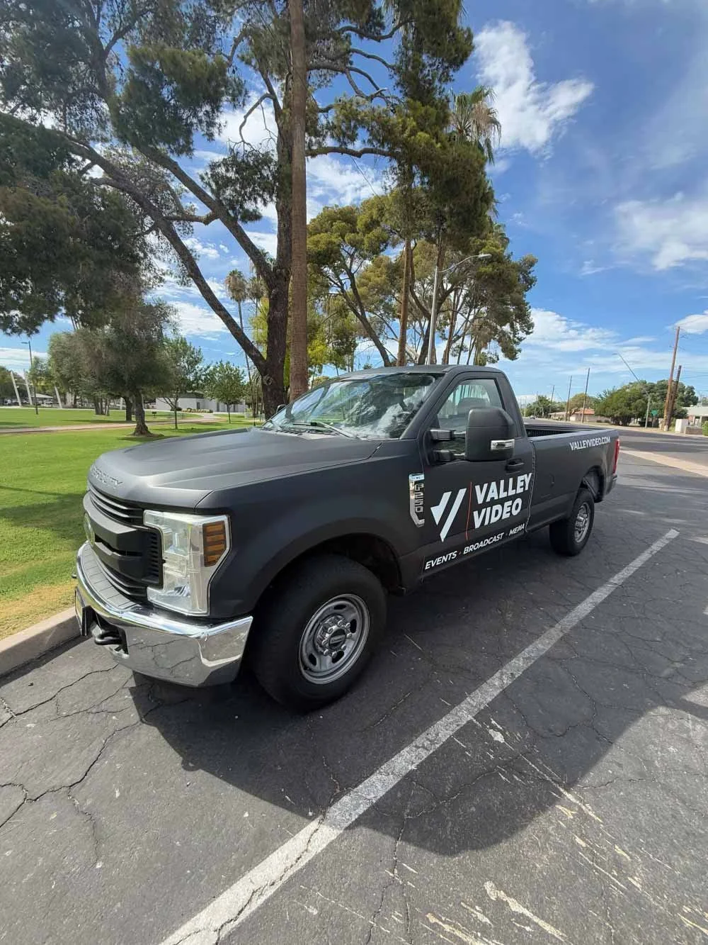 A black pickup truck with Valley Video logos parked on an asphalt street with green grass and tall trees in the background, under a partly cloudy sky.