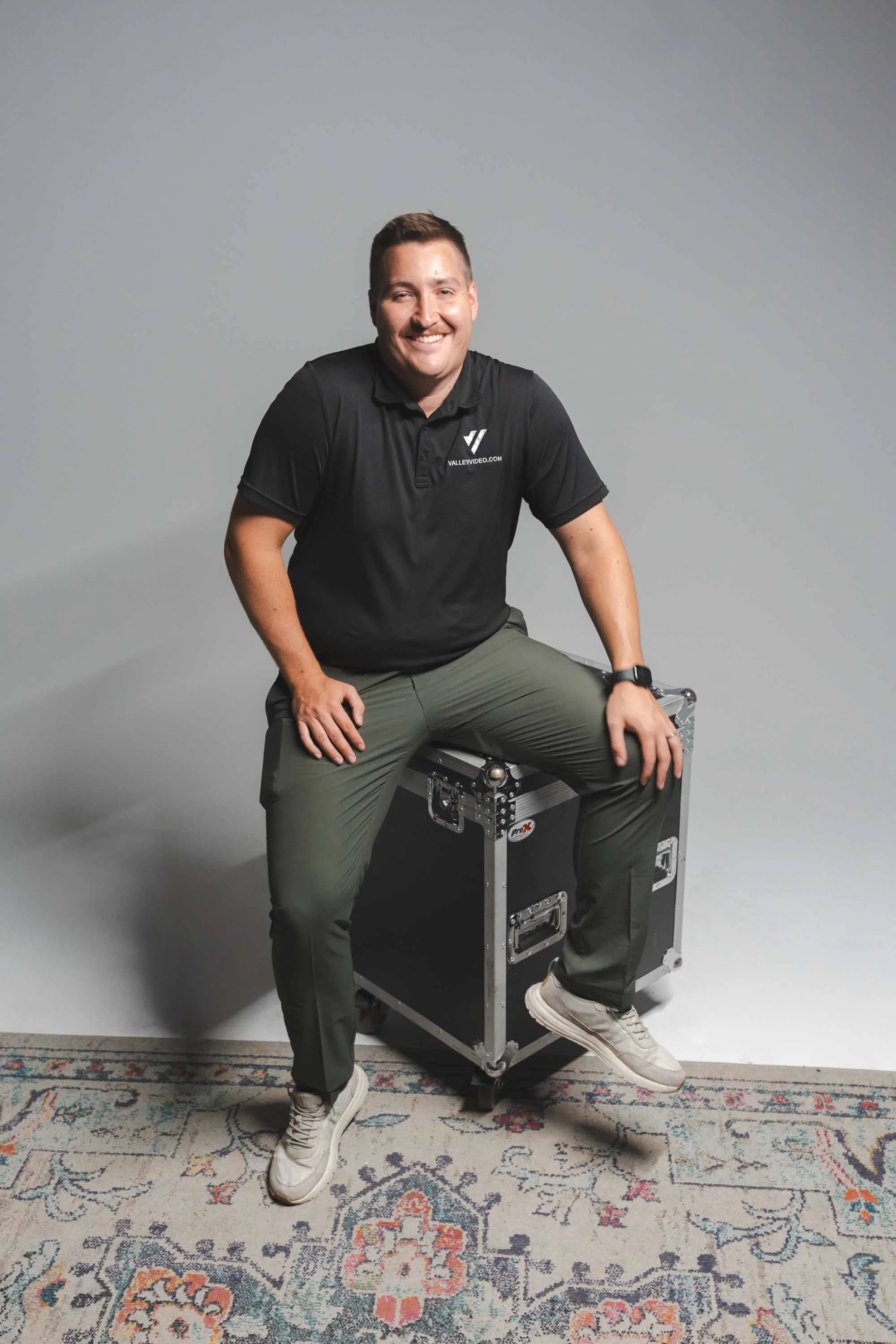 A smiling man wearing a black polo shirt with the Valley Video logo, khaki pants, and white sneakers, sitting on a black equipment case against a plain gray background, with a colorful patterned rug underneath.