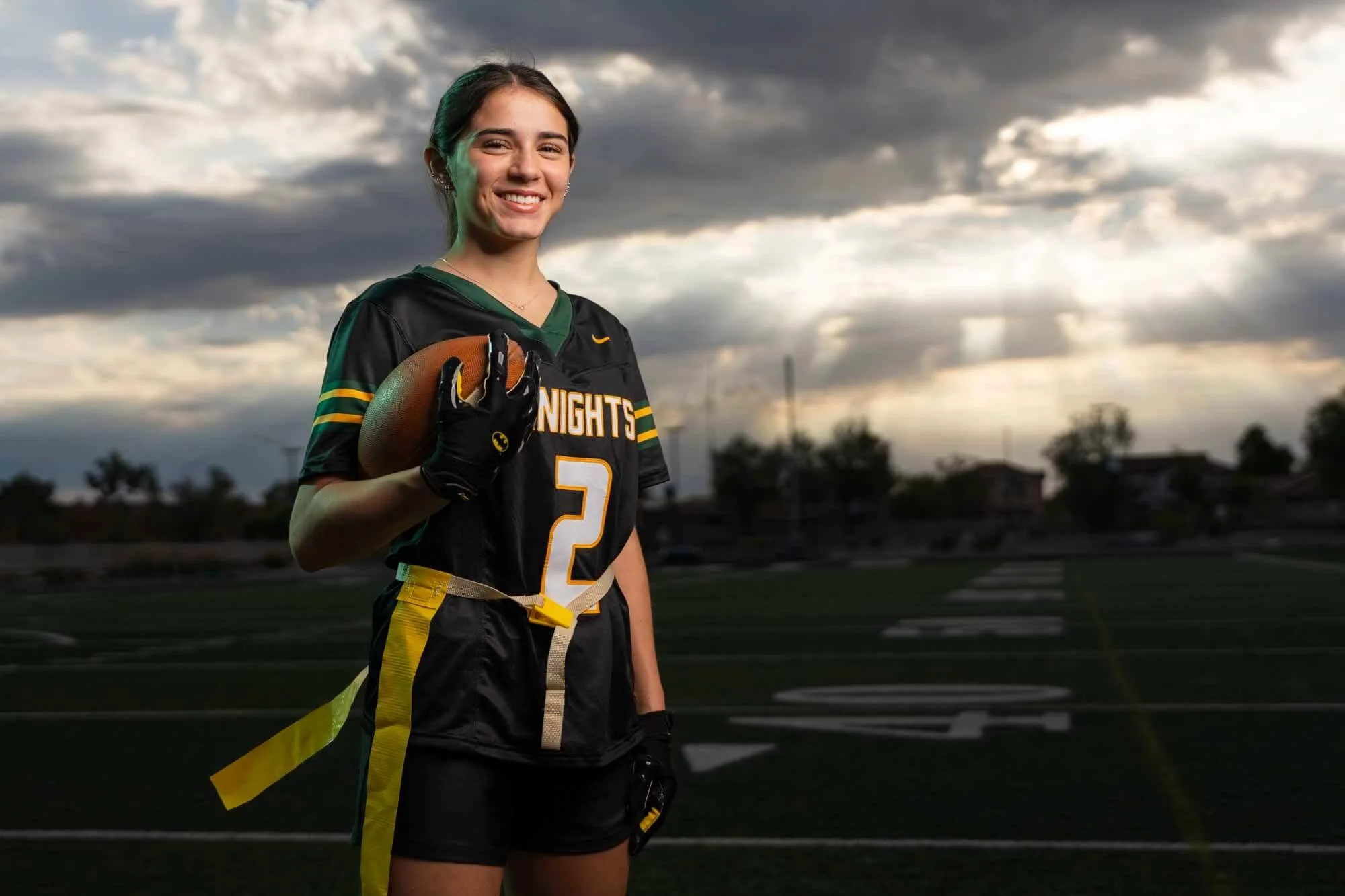 A young female football player standing on a field outdoors during dusk, holding a football, smiling with short dark hair, wearing a black and green football uniform with the number 2, and yellow tape on her waist, under a cloudy sky.