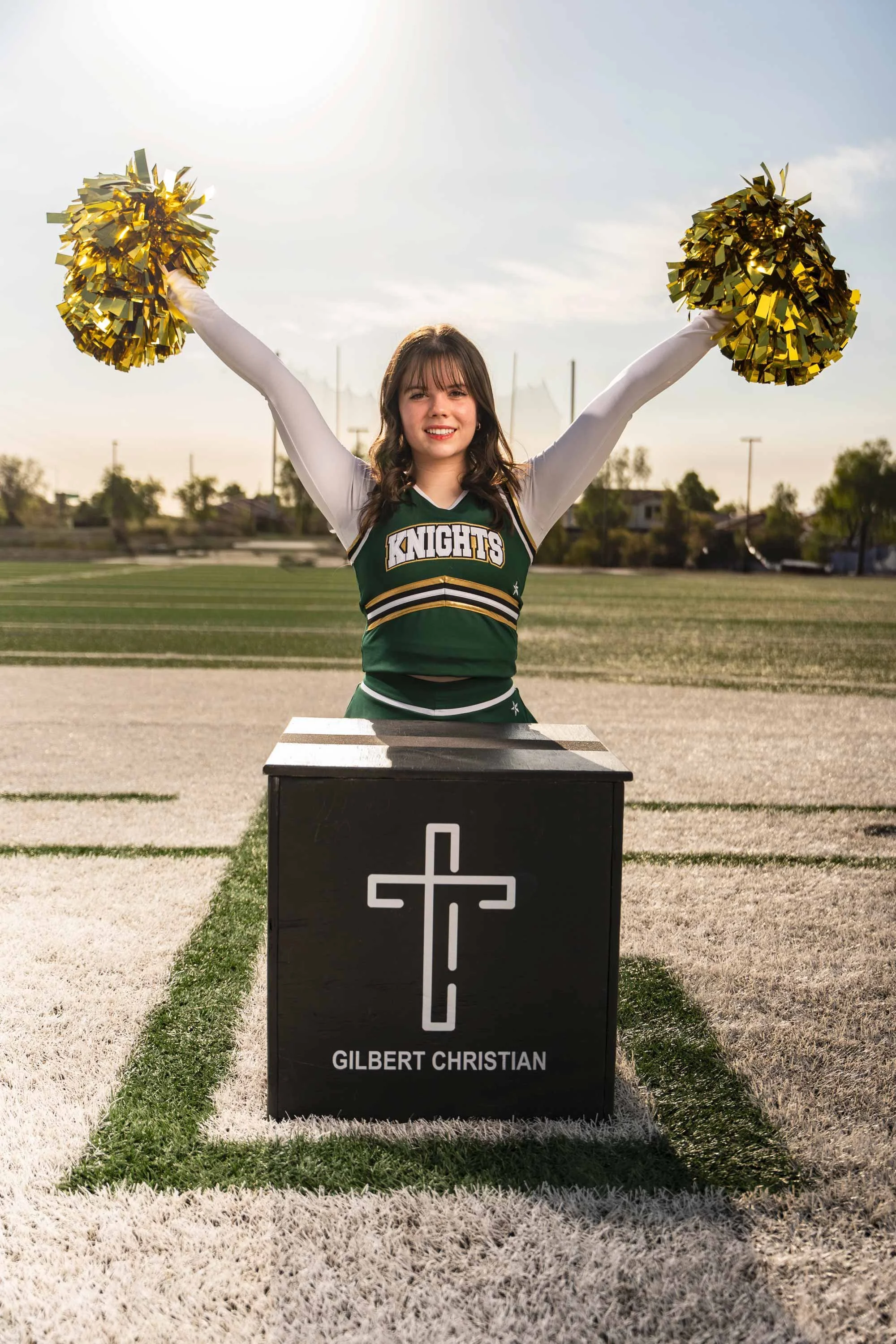A young woman in a cheerleading uniform standing outdoors on a sports field, holding gold pom-poms in the air. There is a black memorial box with a white cross and the name   GILBERT CHRISTIAN in front of her. The sky is partly cloudy and the background includes trees and light poles.