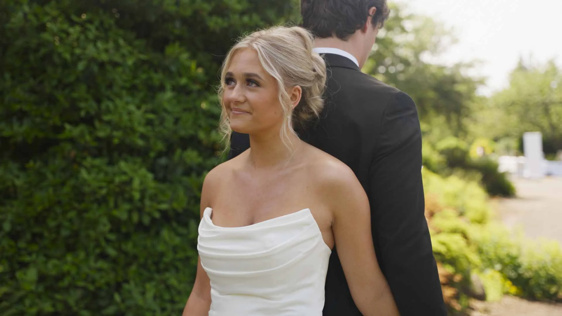Valley Video wedding videography services. A bride in a strapless white wedding dress stands outdoors, smiling softly, with a groom in a black tuxedo back-to-back behind her, against a background of green foliage and trees.
