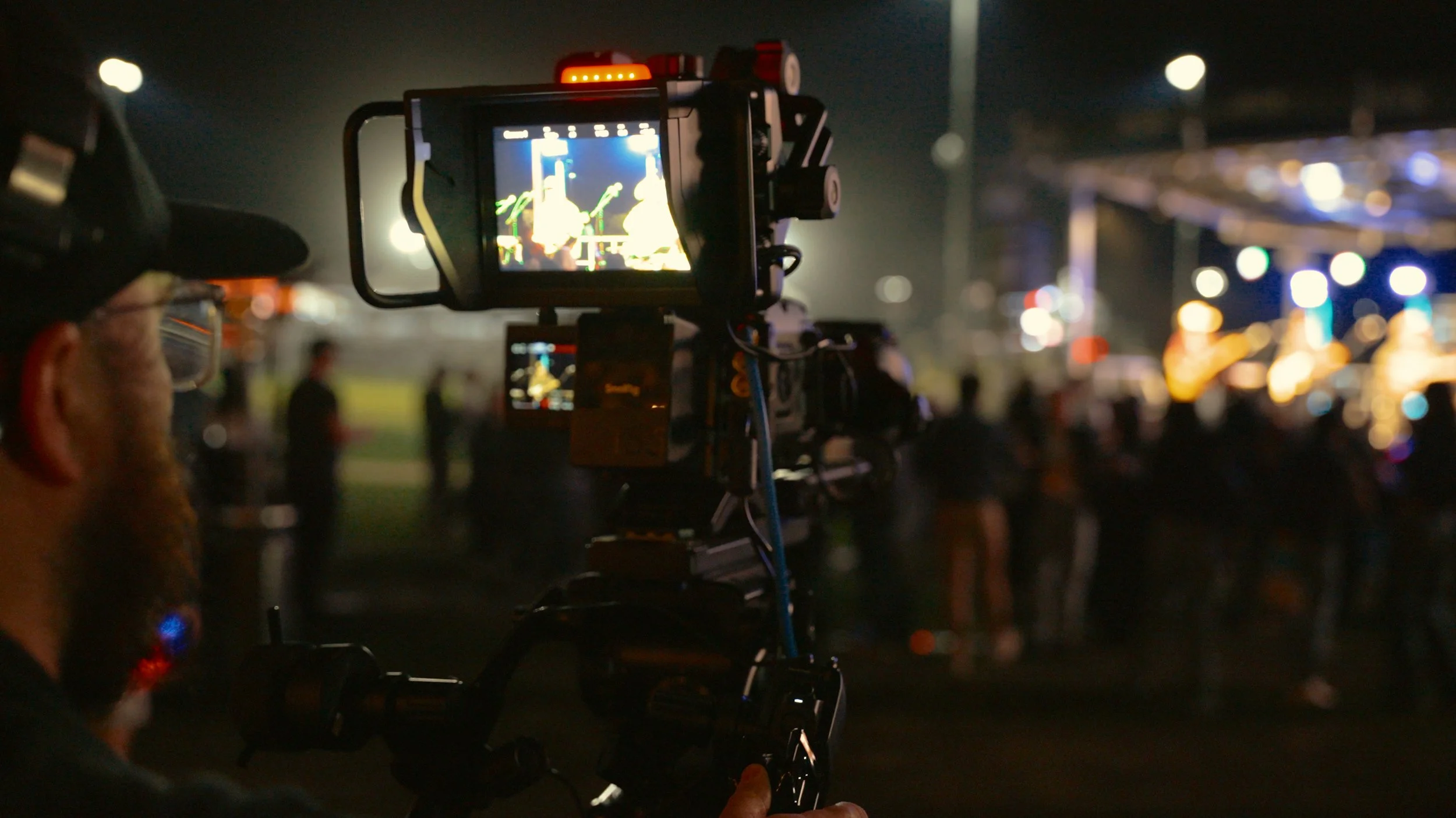 Valley Video behind-the-scenes photo from a production filming event, conference, or concert. A person operating a professional video camera at a nighttime outdoor music concert, with stage lights and a blurred crowd in the background.