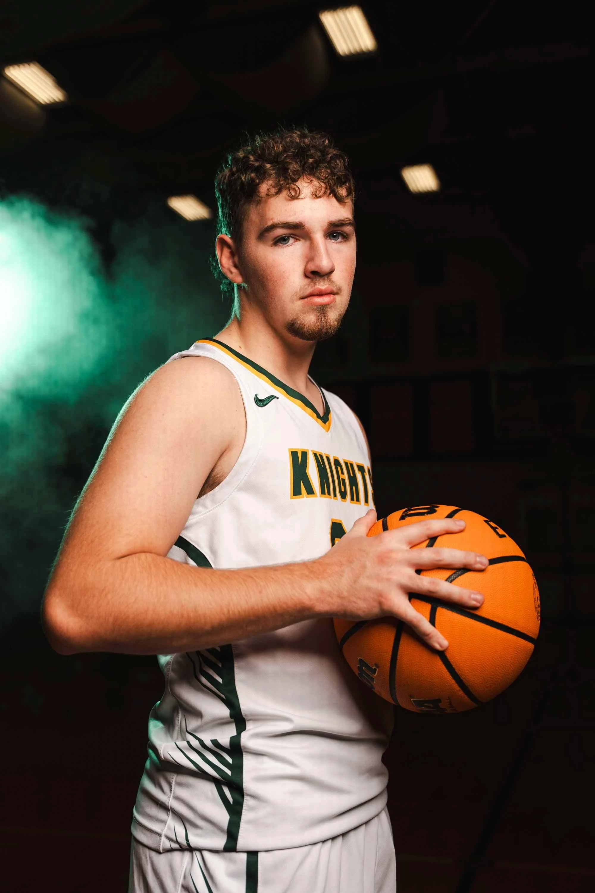 A young male basketball player in a white basketball uniform holding an orange basketball in a dark studio environment with green lighting.
