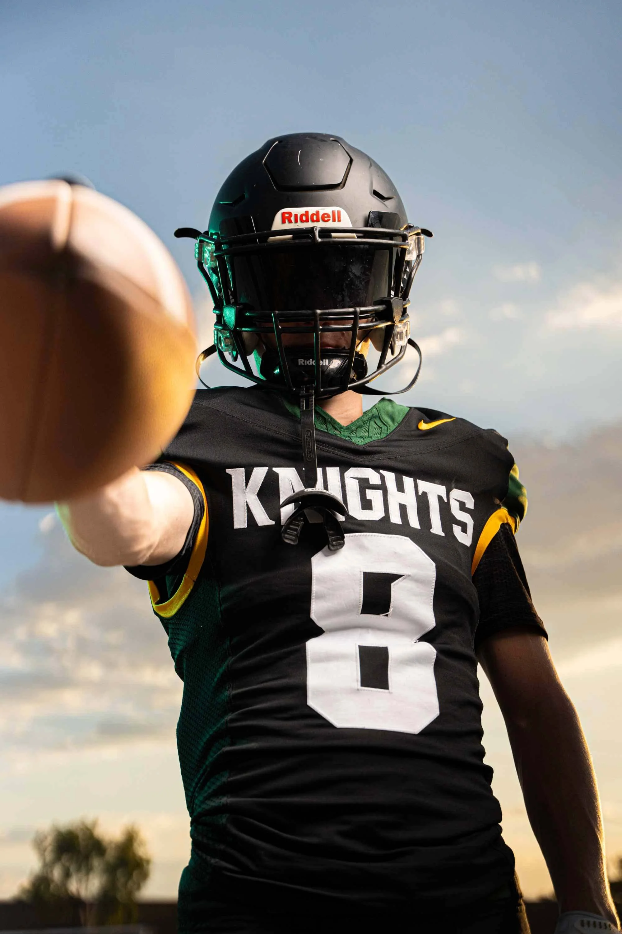 A football player in a black and green uniform with the word 'KNIGHTS' and the number 8 on it, wearing a black helmet with a faceguard, extending his arm toward the camera against a sky background.