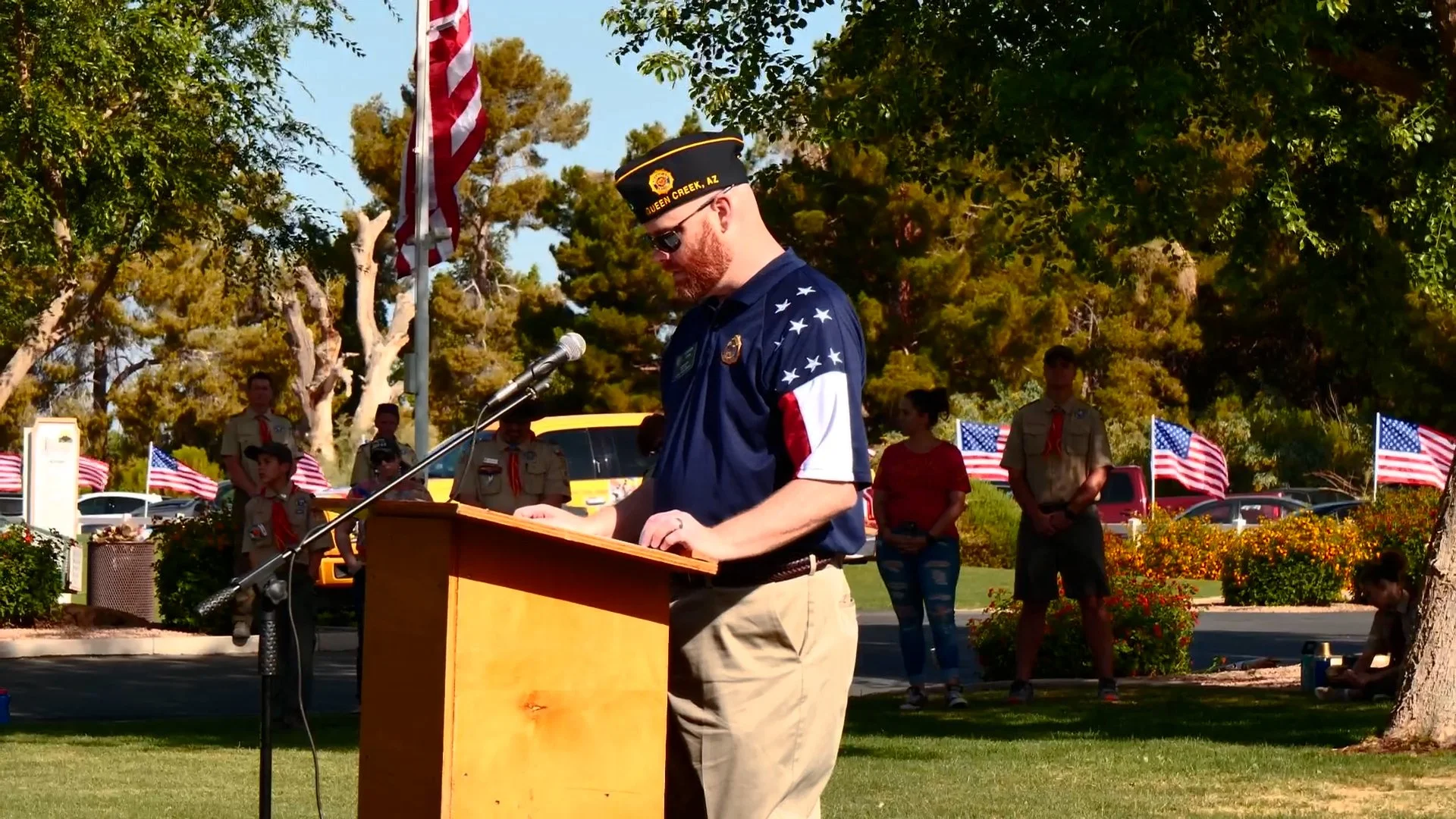 A man in uniform giving a speech at a memorial event, standing behind a wooden podium, with American flags and other people in scouts uniforms and casual clothing in the background, outdoors surrounded by trees.