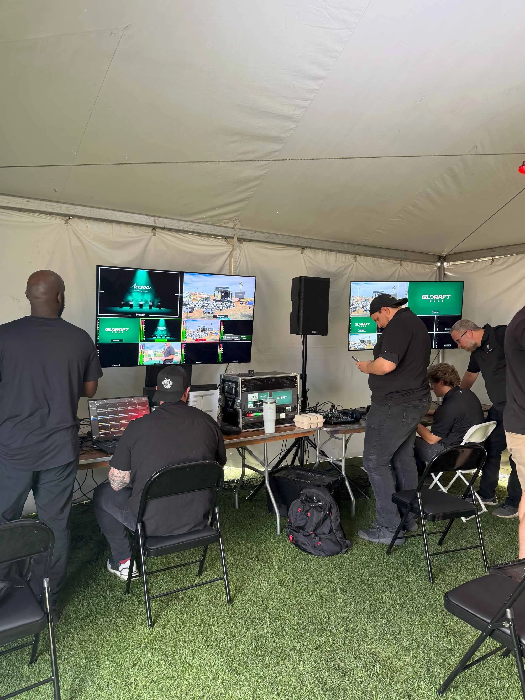 People working on video editing or production setup inside a large white event tent, with multiple monitors, laptops, and professional equipment, during an outdoor event.