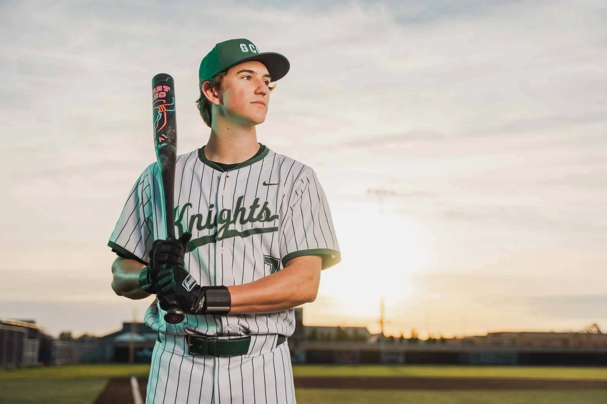 A young baseball player standing on a field during sunset, holding a bat over his shoulder, wearing a gray pinstriped uniform with the word "Knights" on the front and a green cap.