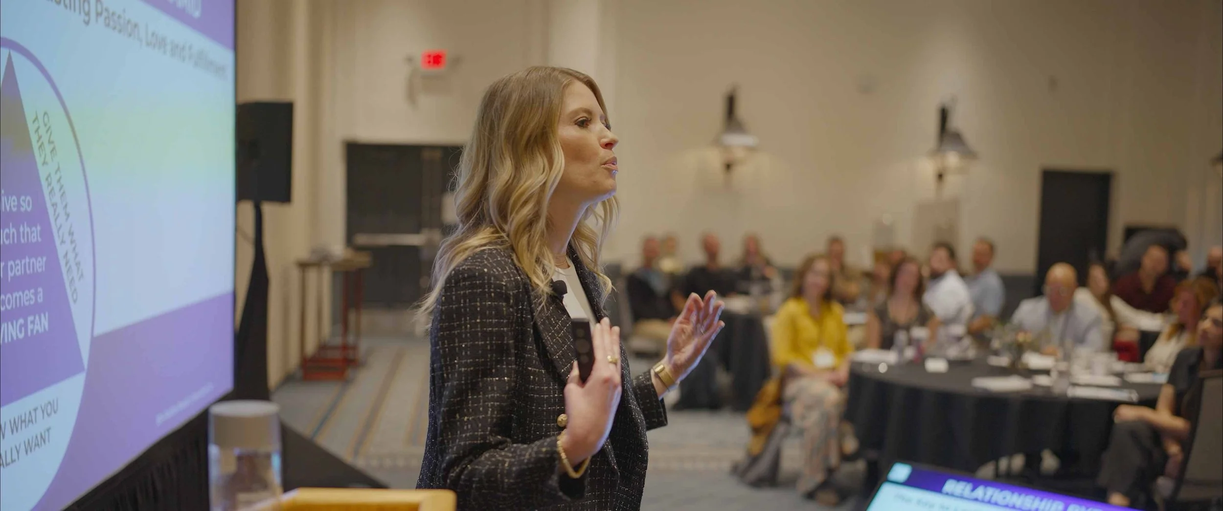 A woman with blonde hair and a checkered blazer gives a presentation in front of a large audience in a conference room. The audience sits at round tables, listening attentively.