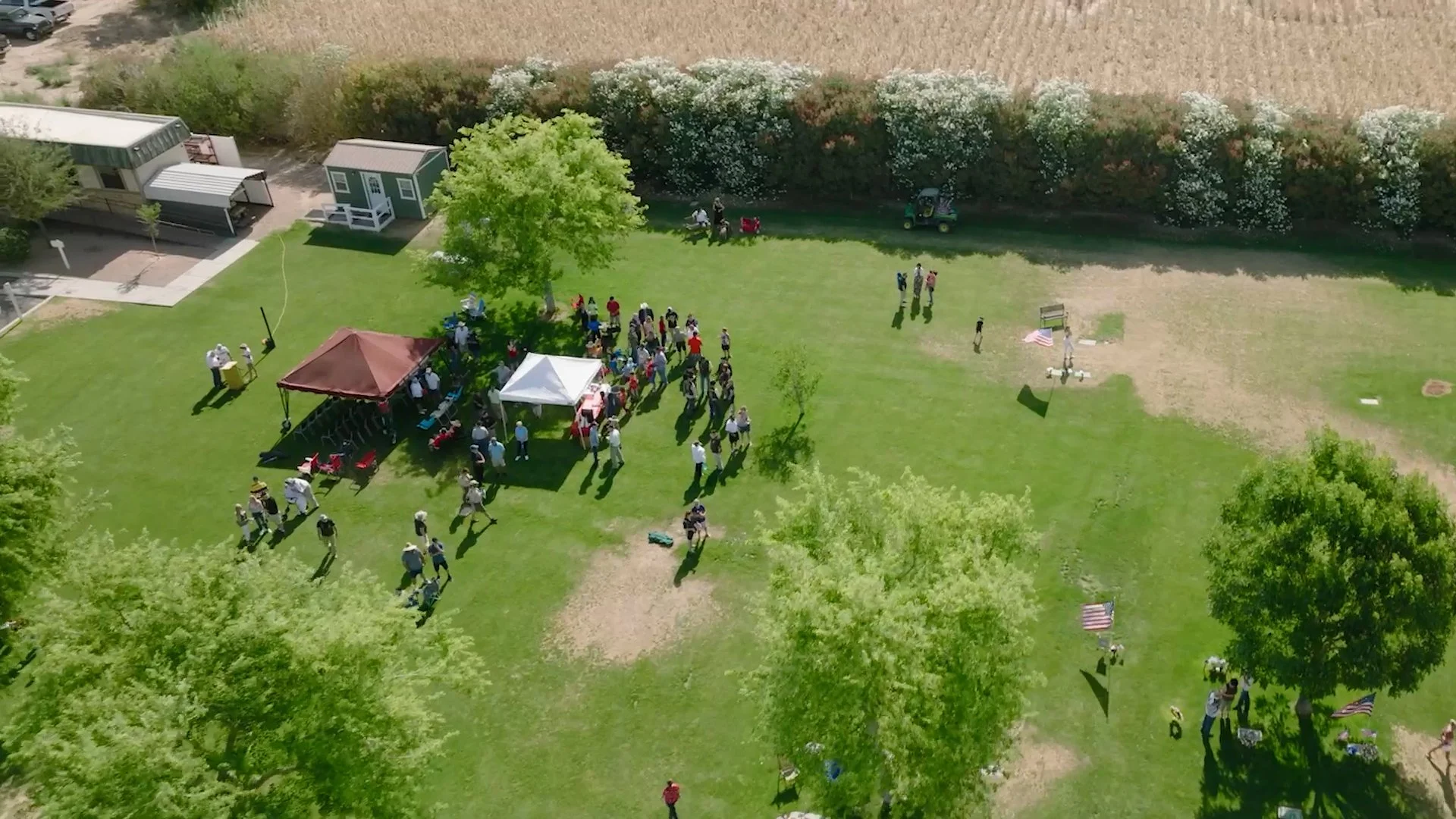 An aerial view of a gathering at a park with a tent, a gazebo, and American flags, including a group of people, some seated and some standing, along with children and adults. There are trees, open grass, and a paved pathway in the park, with a building and a field visible in the background.