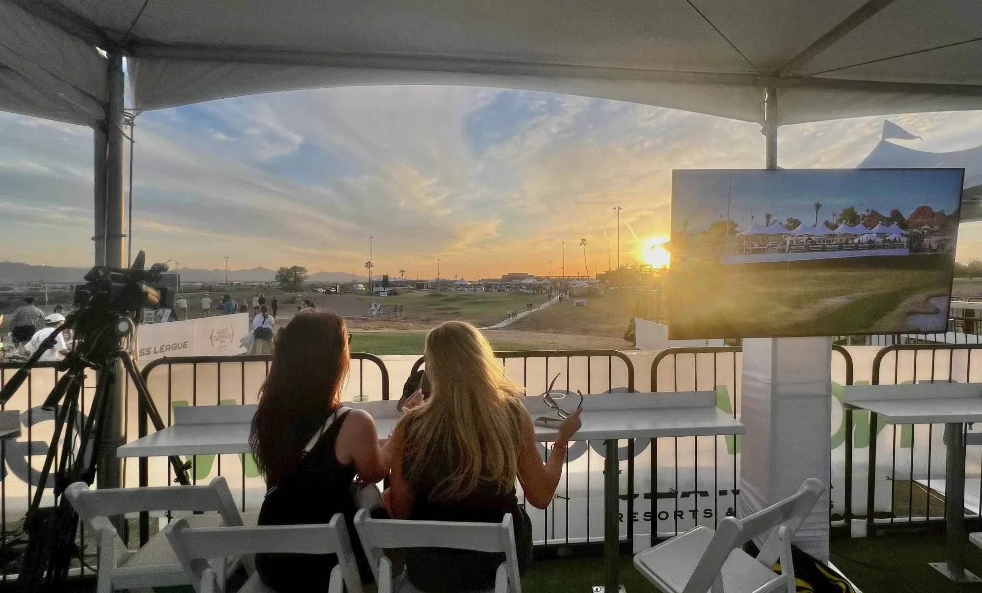 Two women sitting at a table inside a tent, looking at a large screen displaying outdoor scenery with tents and trees, during a sunset at what appears to be a sports or outdoor event.