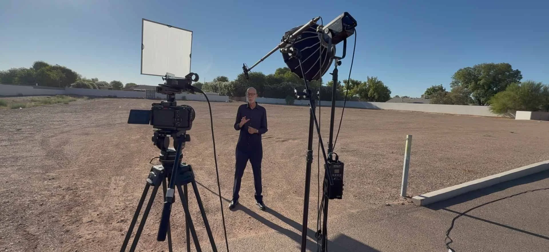 A man in black clothes stands in an empty lot, surrounded by professional video and photography equipment, under a clear blue sky.