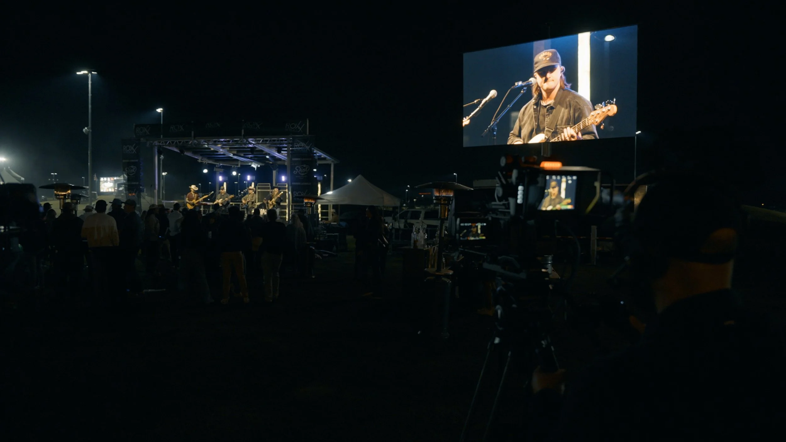 Nighttime outdoor concert with a band performing on a stage, large screen showing a musician playing guitar, audience gathered in front, some people under umbrellas, various equipment and lighting around.