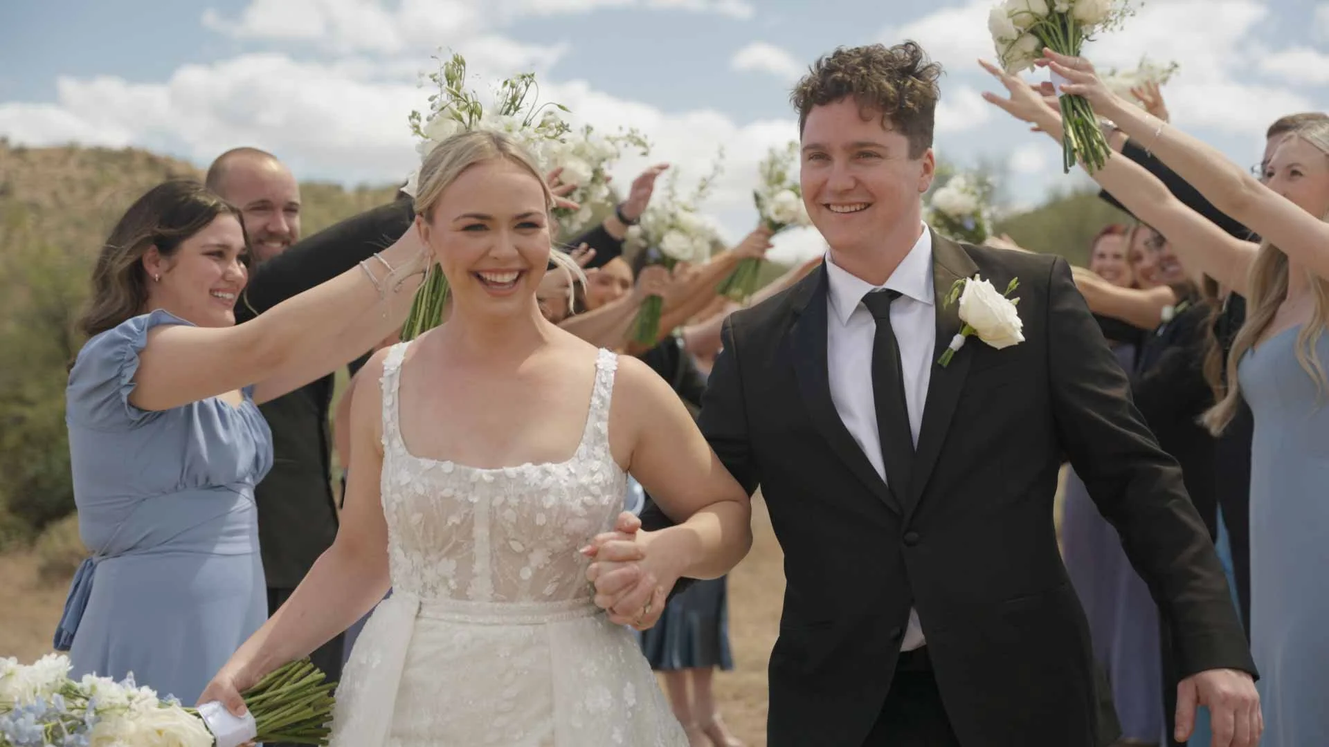 Valley Video wedding videography services. Bride and groom holding hands walking outdoors, surrounded by friends holding bouquets, celebrating wedding outdoors with cloudy sky and landscape in background.