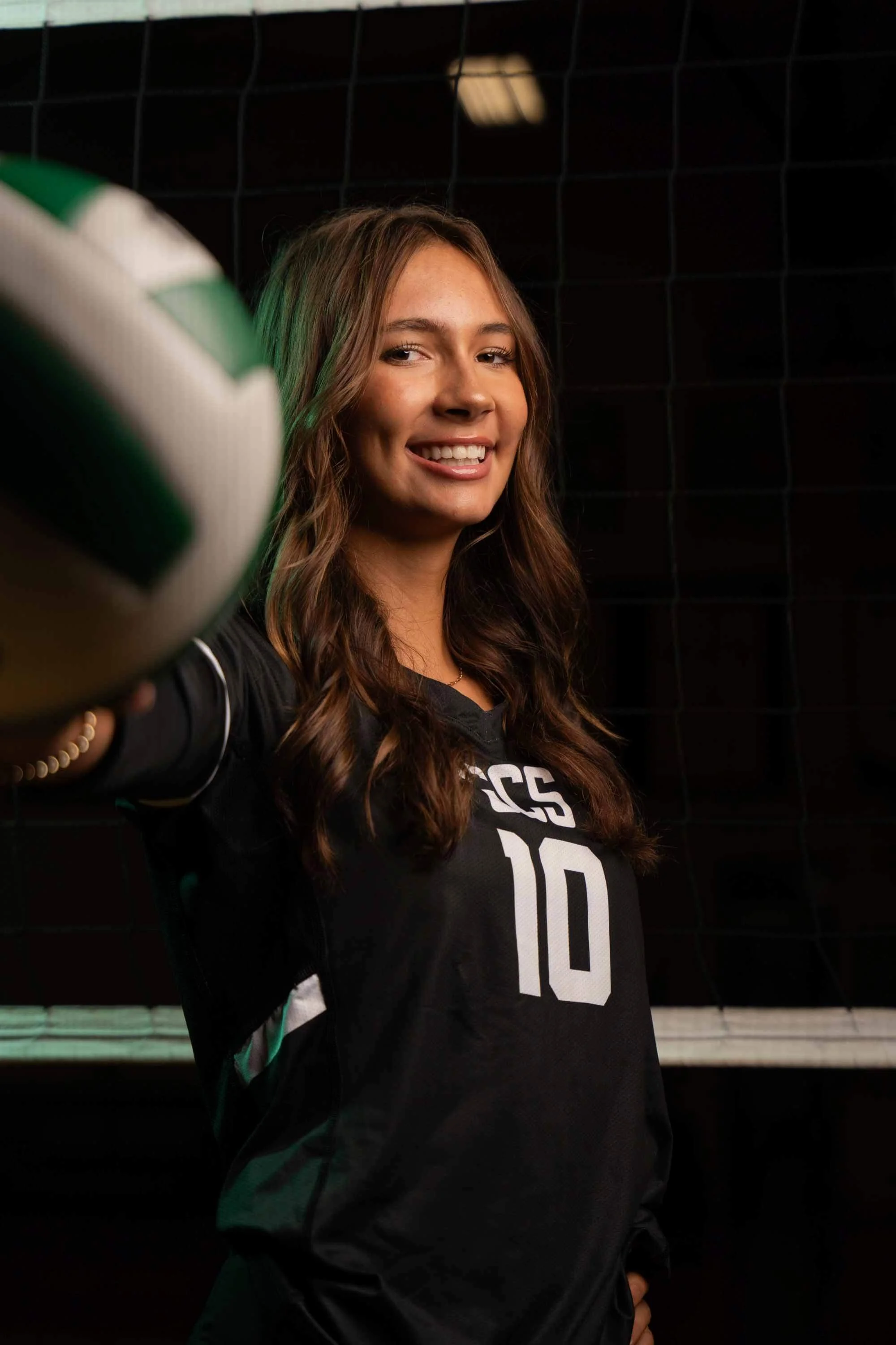 A young woman with long wavy brown hair smiling and holding a green and white volleyball. She is wearing a black sports jersey with the number 10 on it. The background is dark with a black net and some lighting overhead.