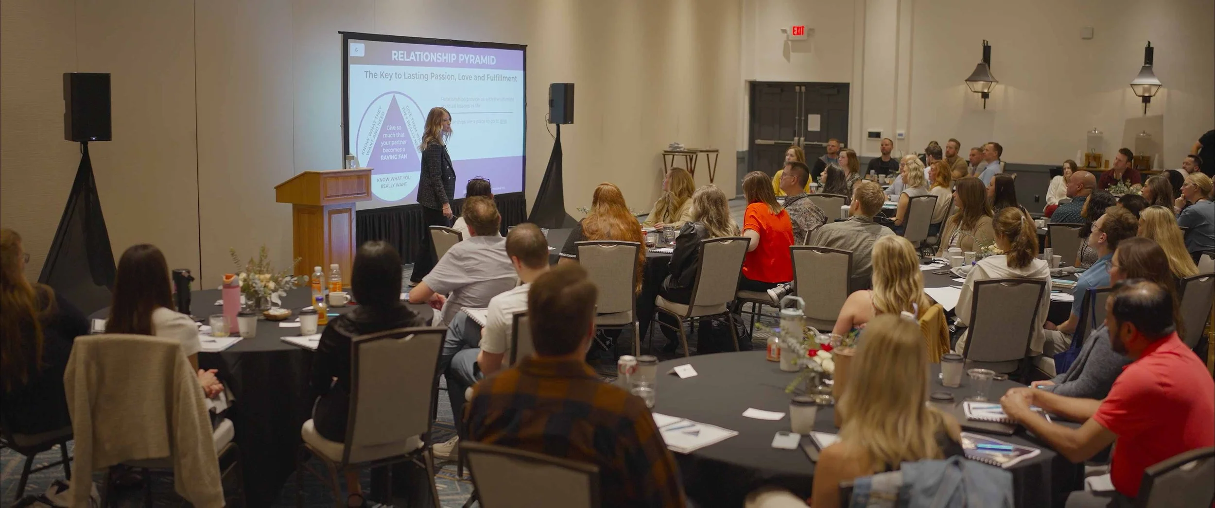 A woman is giving a presentation to a large audience in a conference room. The presentation slide behind her reads 'Relationship Pyramid' and contains text about passion, love, and fulfillment. The room is filled with round tables of attendees, some taking notes, and decorated with flowers and drinks.