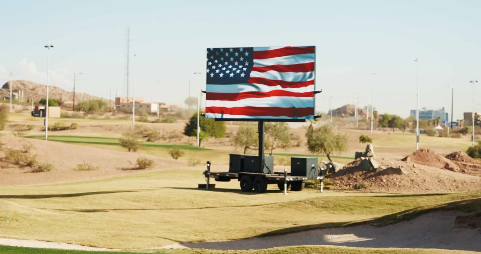 Large American flag billboard on a trailer in a desert-like area, with a golf course in the foreground and hills and buildings in the background.