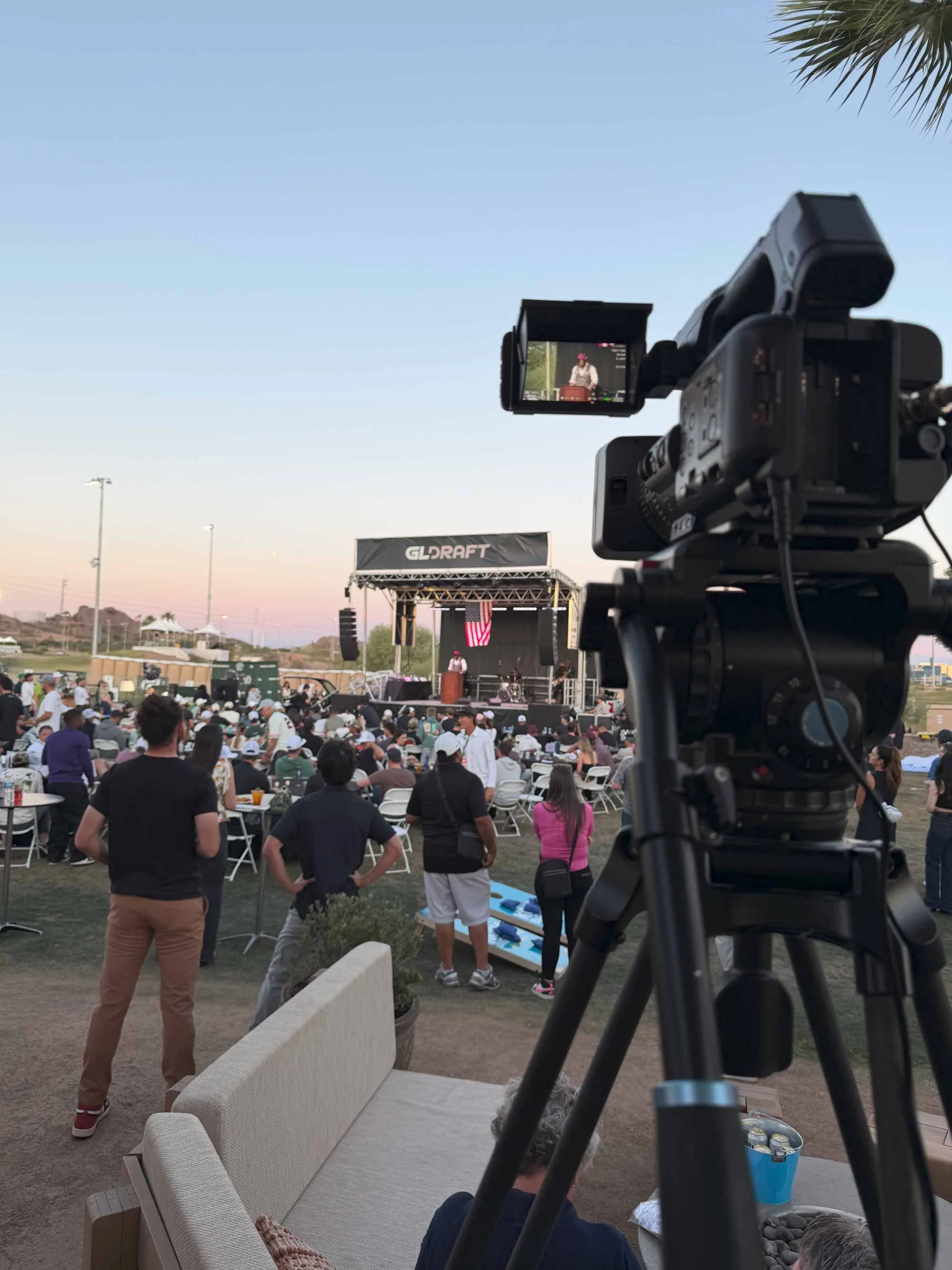 A daytime outdoor event with a crowd of people seated at tables, a stage with a person speaking at a podium, and video recording equipment capturing the scene. There is a large screen and an American flag visible on the stage.