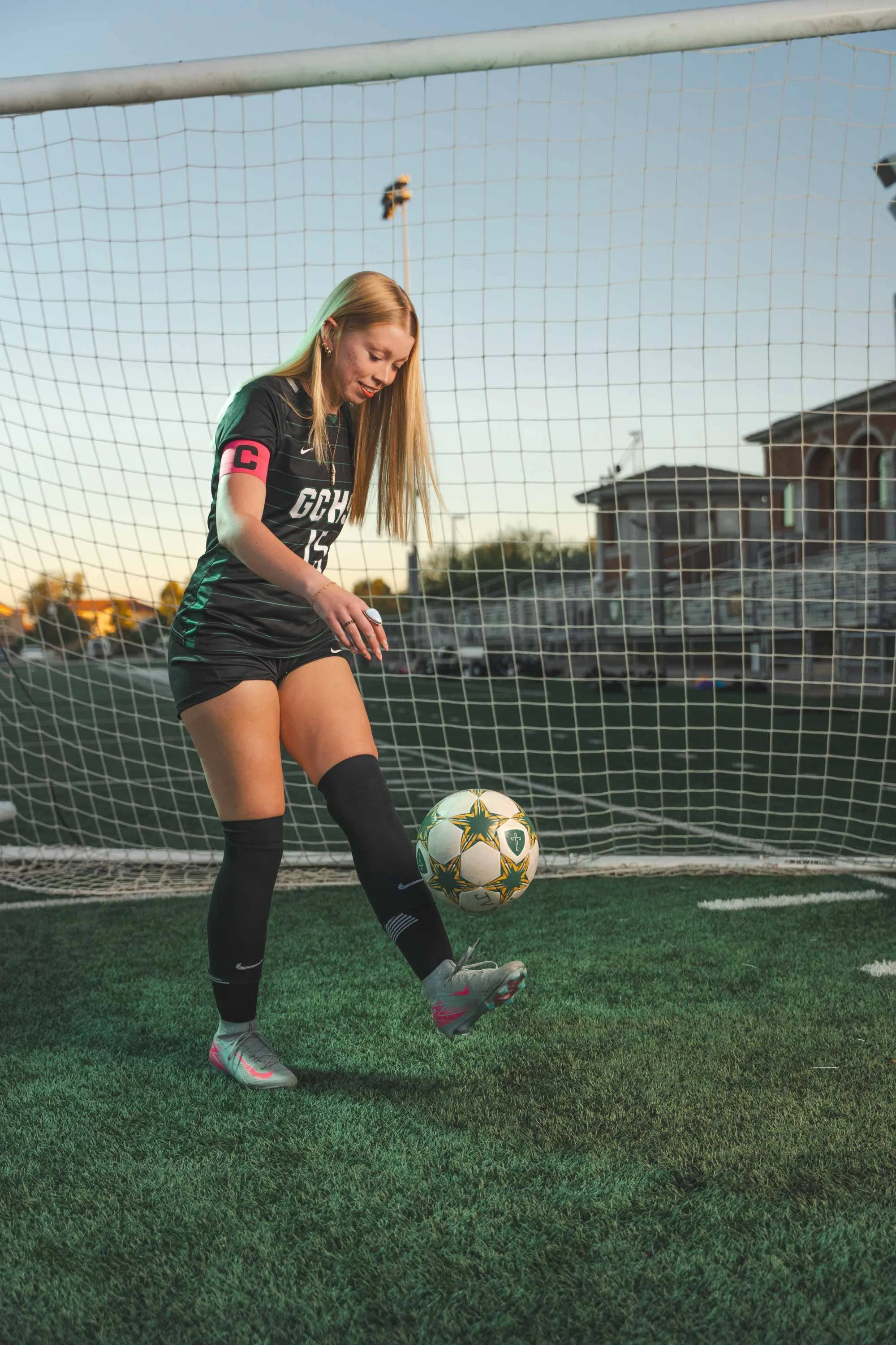 A female soccer player in a black uniform with a Captain's armband is on a soccer field, about to kick a soccer ball. The background shows a goal net, houses, and a clear sky during sunset.