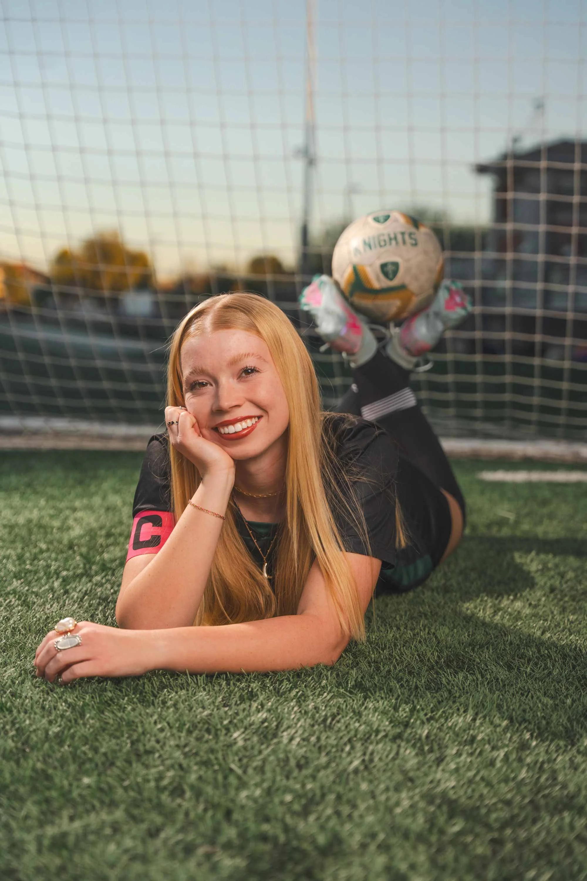 A red-haired woman lying on the grass in front of a soccer goal, smiling at the camera, wearing a black and pink sports jersey, and holding her chin with one hand.