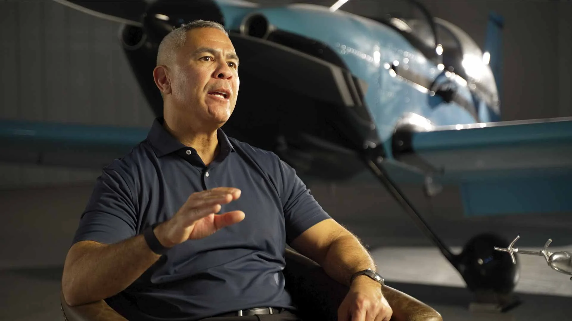 Valley Video interview and talking head video production services example. A man sitting in a chair in front of an aircraft, possibly an airplane or jet, in a hangar or aircraft storage facility.