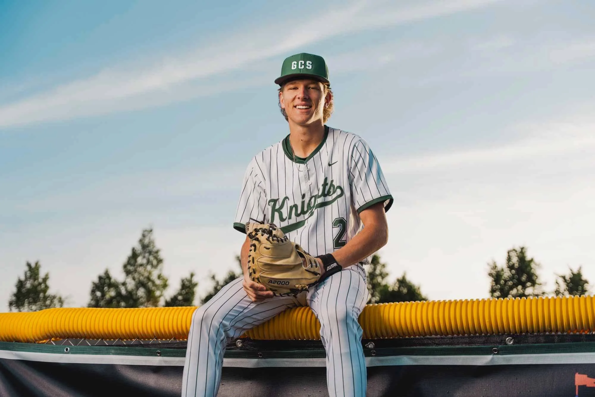 A young baseball player in a pinstripe uniform with 'Knights' written on it, sitting on a yellow barrier, holding a baseball glove, wearing a green cap with 'GCS' on it, outdoors with trees and a cloudy sky in the background.