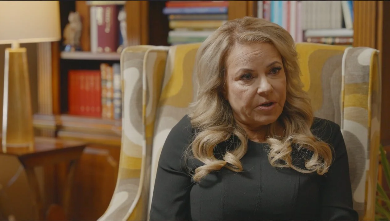 Valley Video interview and talking head video production services example. A woman with blonde hair seated in a patterned armchair in a room with bookshelves, reading lamp, and decorative items.