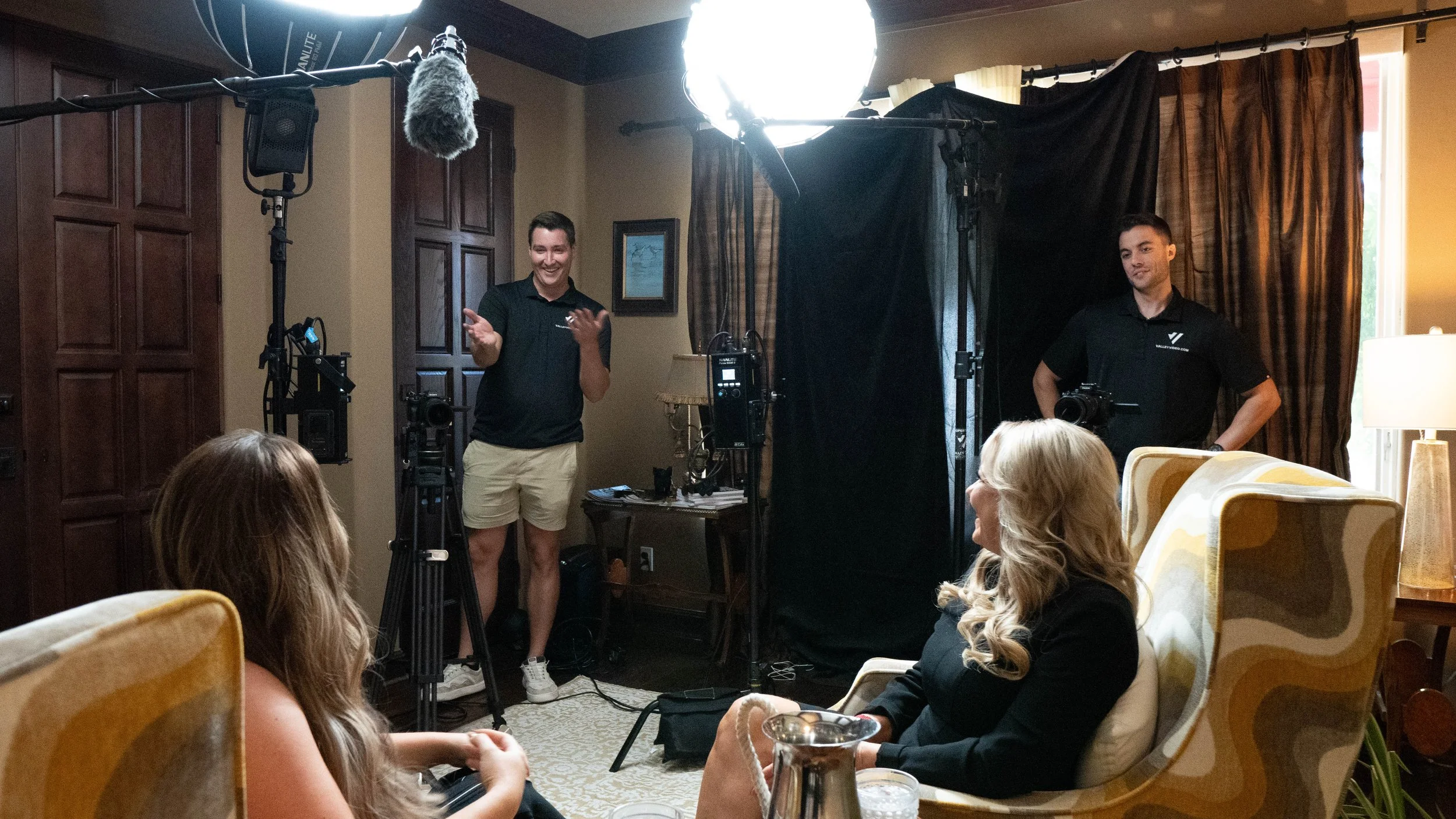The Valley Video crew filming an interview or a presentation in a cozy, warmly lit room with two women seated on patterned armchairs and two men standing in front of a black screen backdrop surrounded by film equipment.