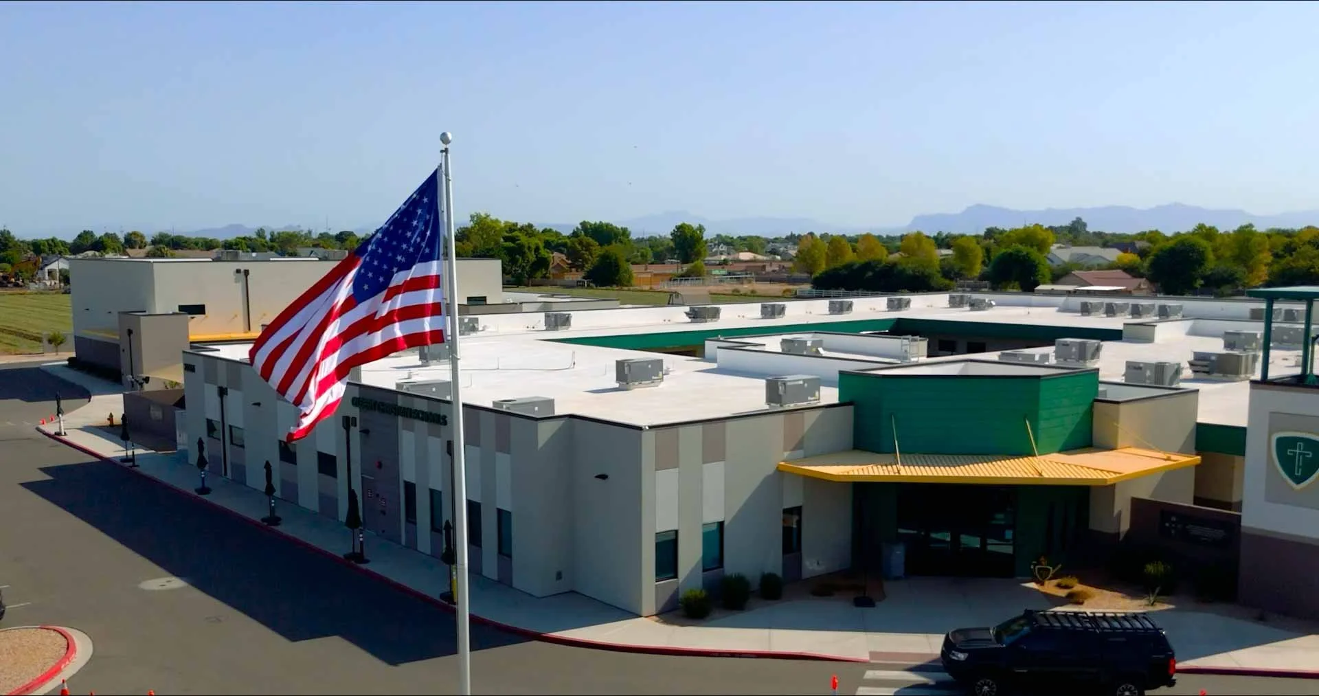An American flag flying in front of a modern building with a flat roof, surrounded by parking lot and greenery, with mountains in the background.