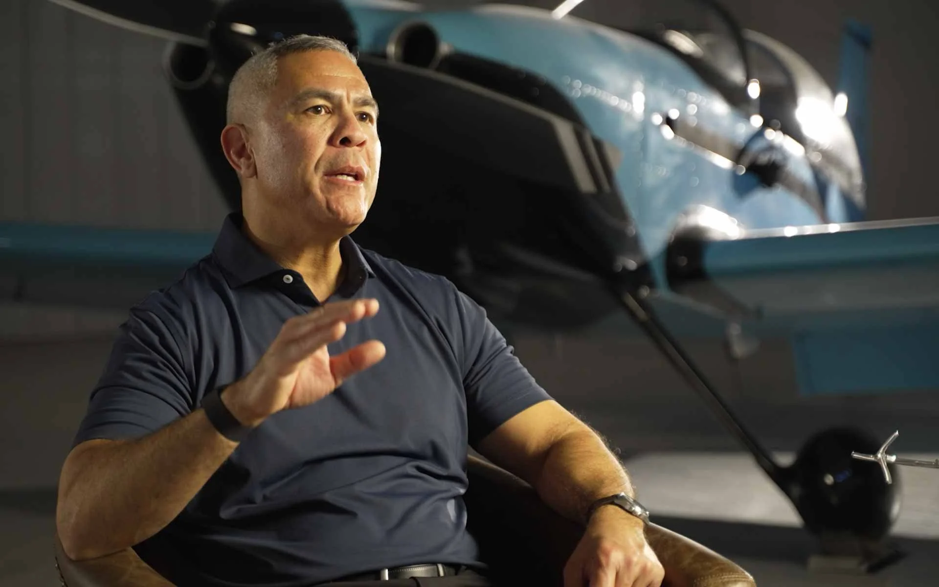 A man with short gray hair wearing a navy blue polo shirt is sitting in a chair gesturing with his right hand, in front of a helicopter.