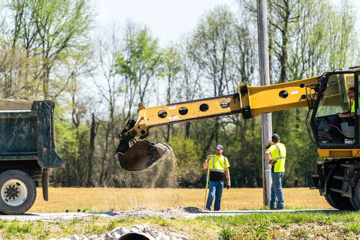 Construction workers and machinery installing a utility pole outdoors on a sunny day, with trees in the background.