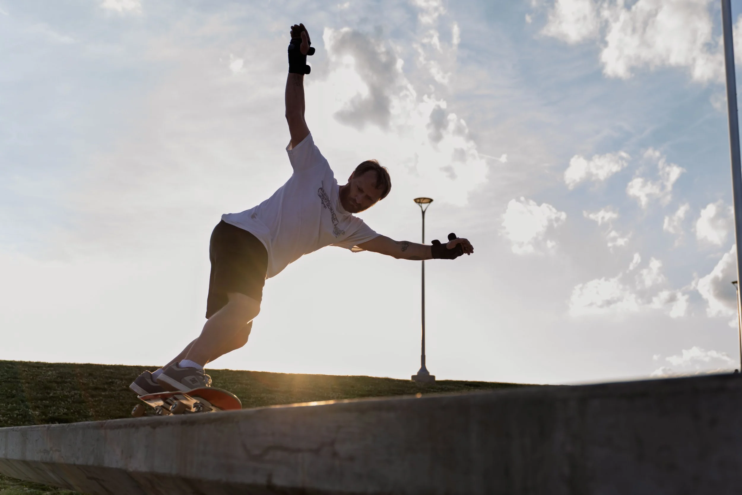 A man skateboarding outdoors in the evening with the sun setting behind him, sky partly cloudy, and street lamps visible in the background.