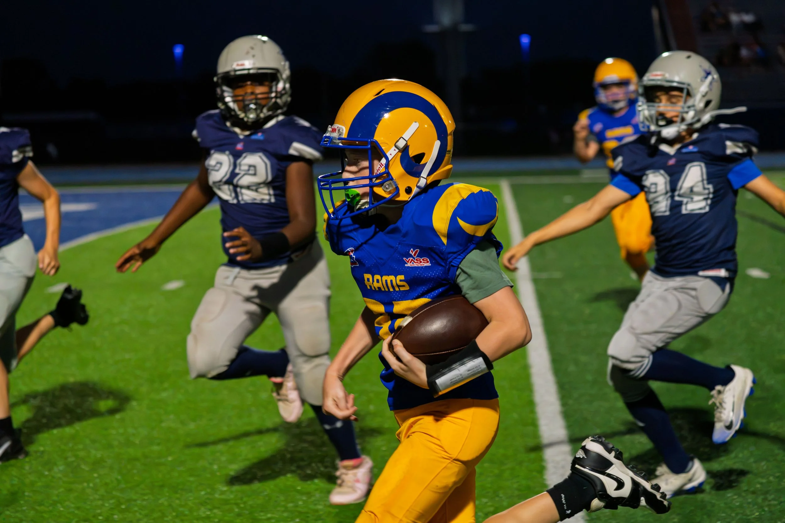 Young football players in blue and yellow uniforms playing on a field during night time. One player in yellow and blue gear holds the football while other players chase him.