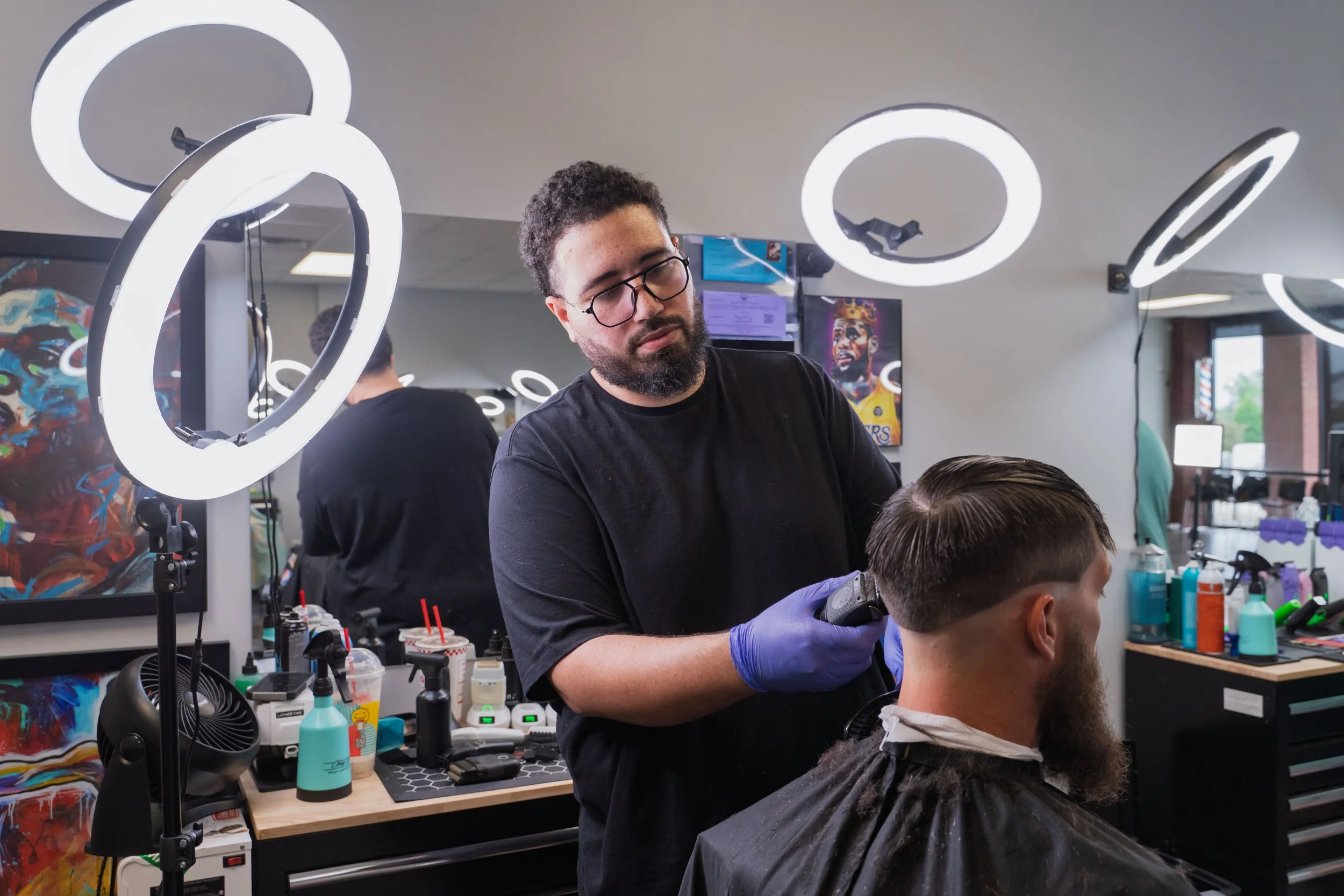 A hairstylist with glasses and a beard cutting a young man's hair in a barbershop. The barbershop has bright lighting, with ring lights, colorful artwork, and various hair styling tools and products on the counter.