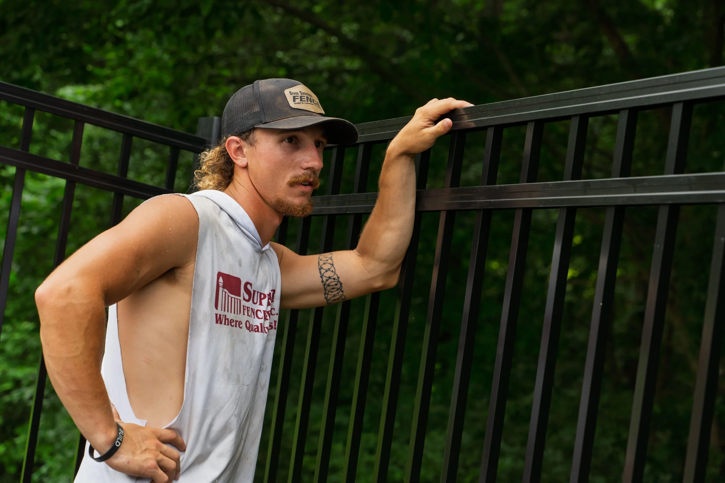 A man with curly hair, a beard, and a tattoo on his arm, wearing a gray cap and a sleeveless shirt, is leaning on and holding a black metal fence with one hand in a wooded outdoor setting.