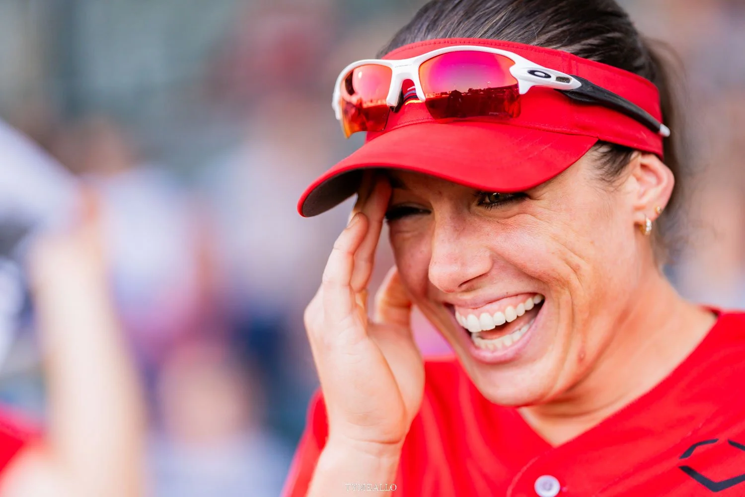 A woman wearing a red cap and sunglasses with a red sports shirt, smiling and touching her forehead.