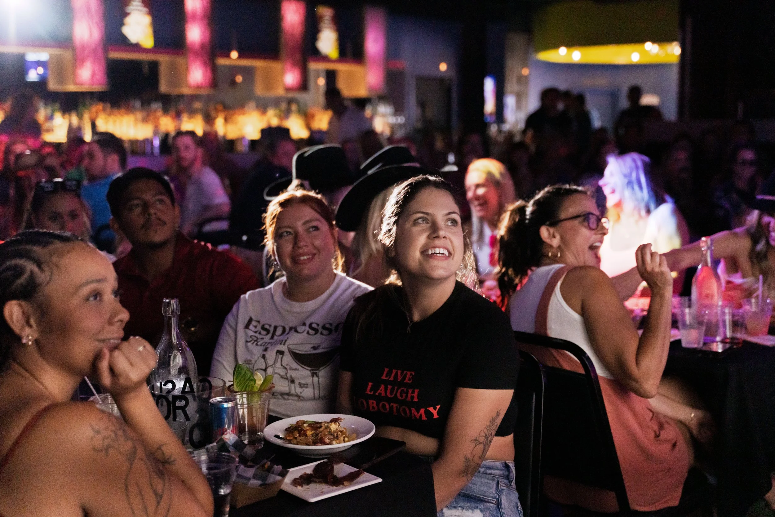 A group of people sitting at a table in a dimly lit nightclub or bar, enjoying food and drinks, with others in the background engaged in conversations and entertainment.