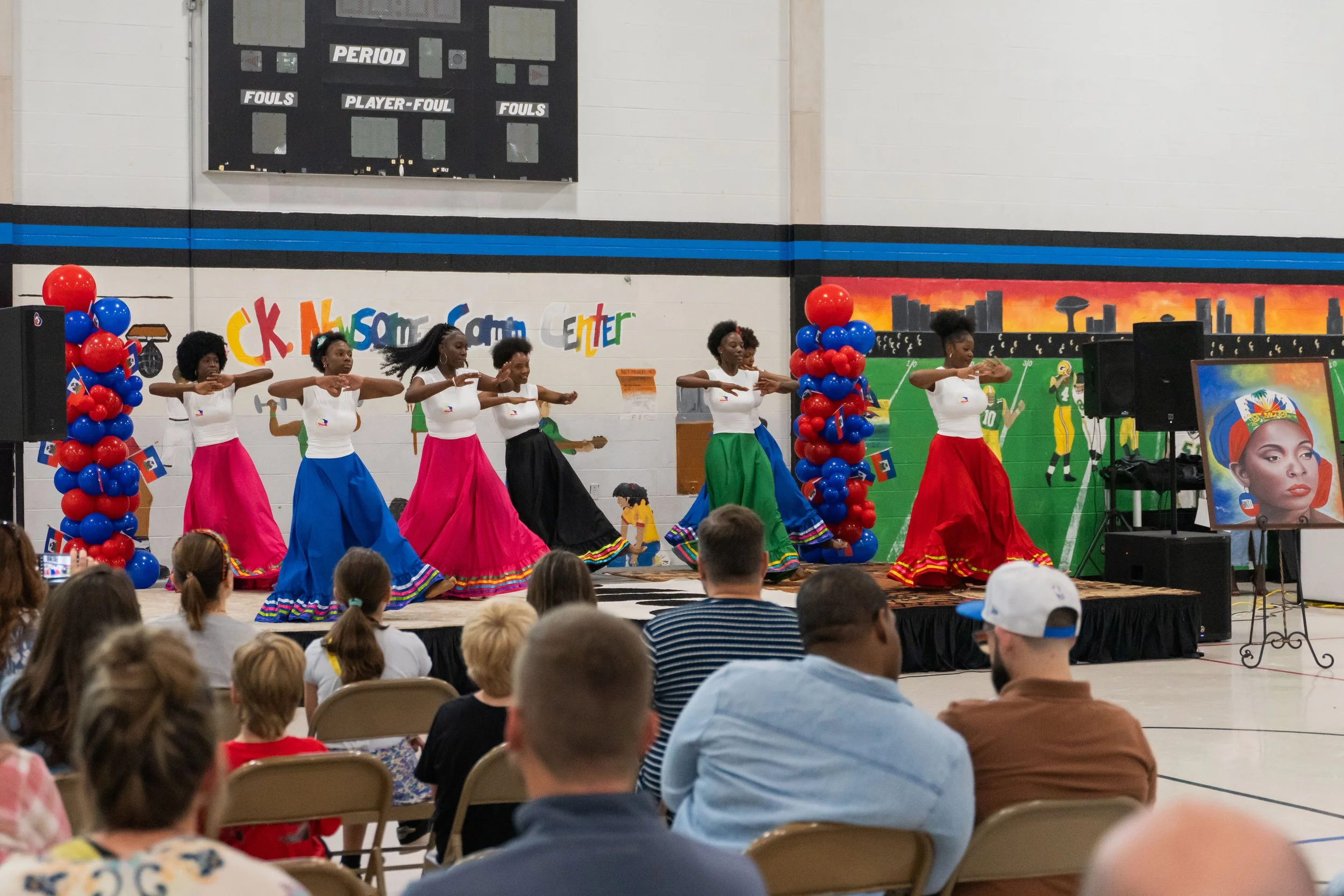 Group of women in colorful skirts and white tops performing a dance on stage with balloons and decorations in a gymnasium setting.