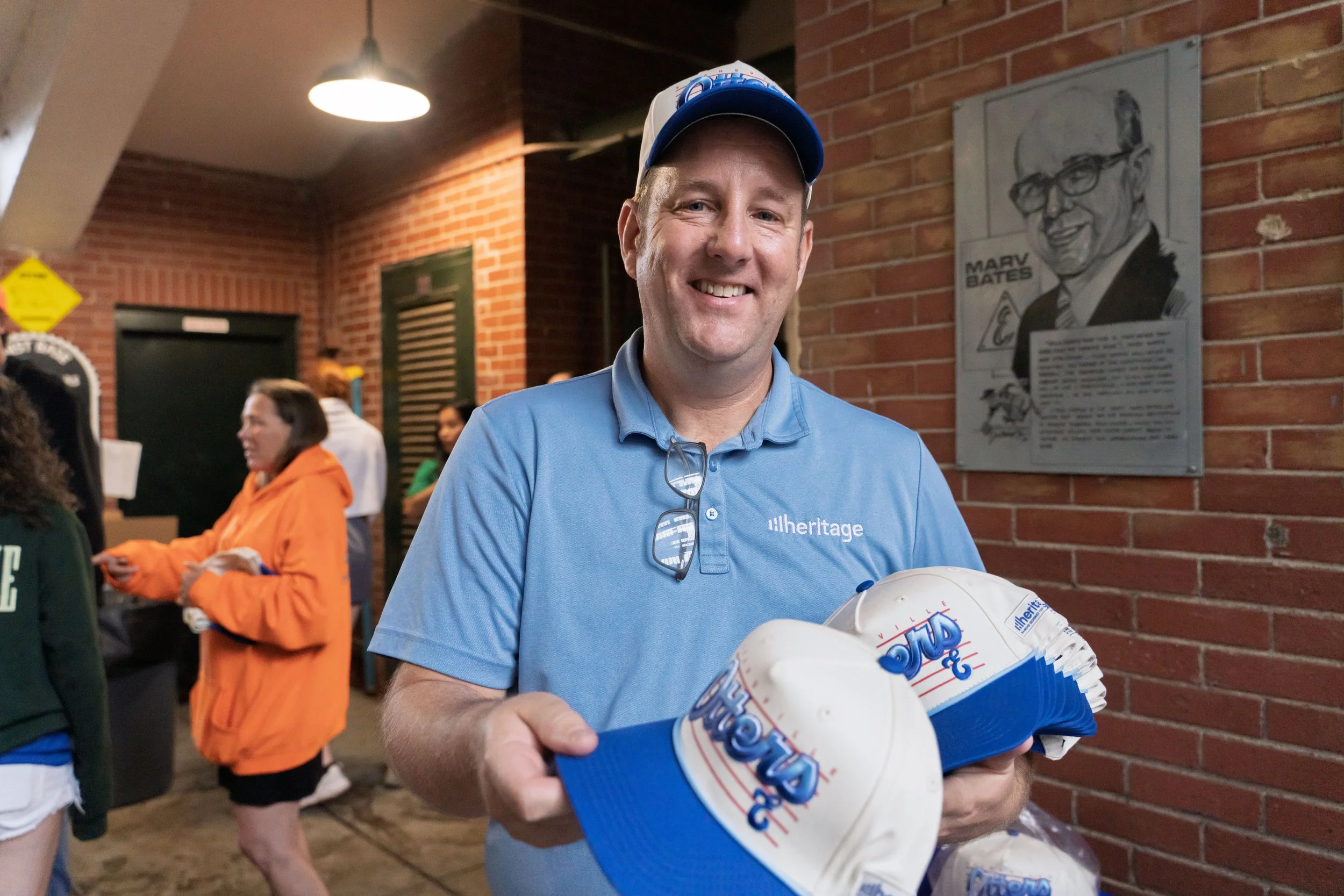 A man in a blue shirt and cap smiling while holding multiple containers of ice cream cones, surrounded by others in a brick-walled indoor setting.