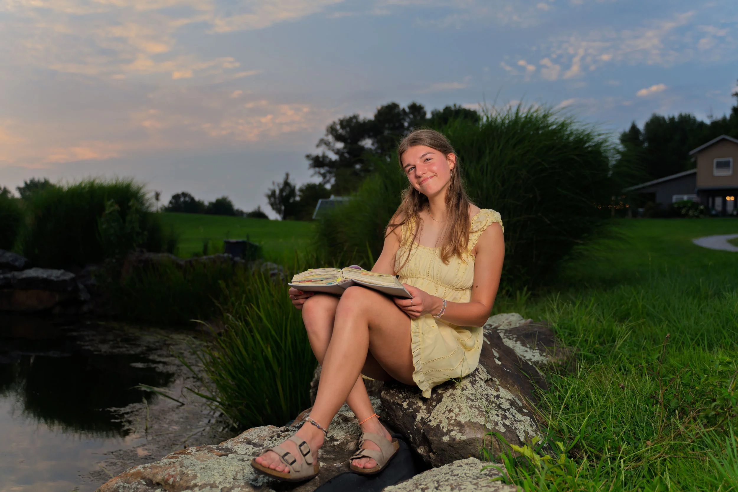 Young girl sitting on a rock by a small stream, holding an open book, smiling at the camera, wearing a yellow dress and sandals, surrounded by greenery with houses in the background during dusk.