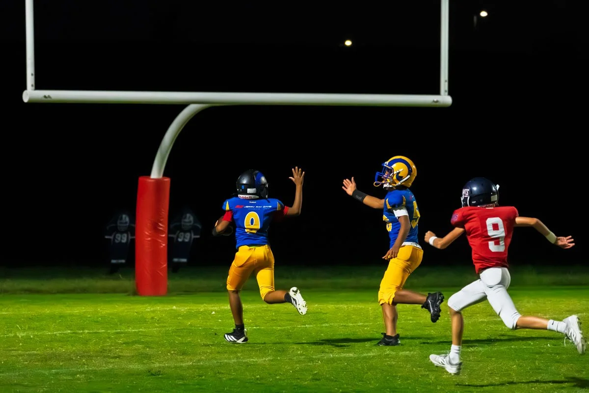 Three young football players on a field at night, one in a red jersey running toward the end zone, and two in blue and yellow jerseys celebrating near the goalpost.