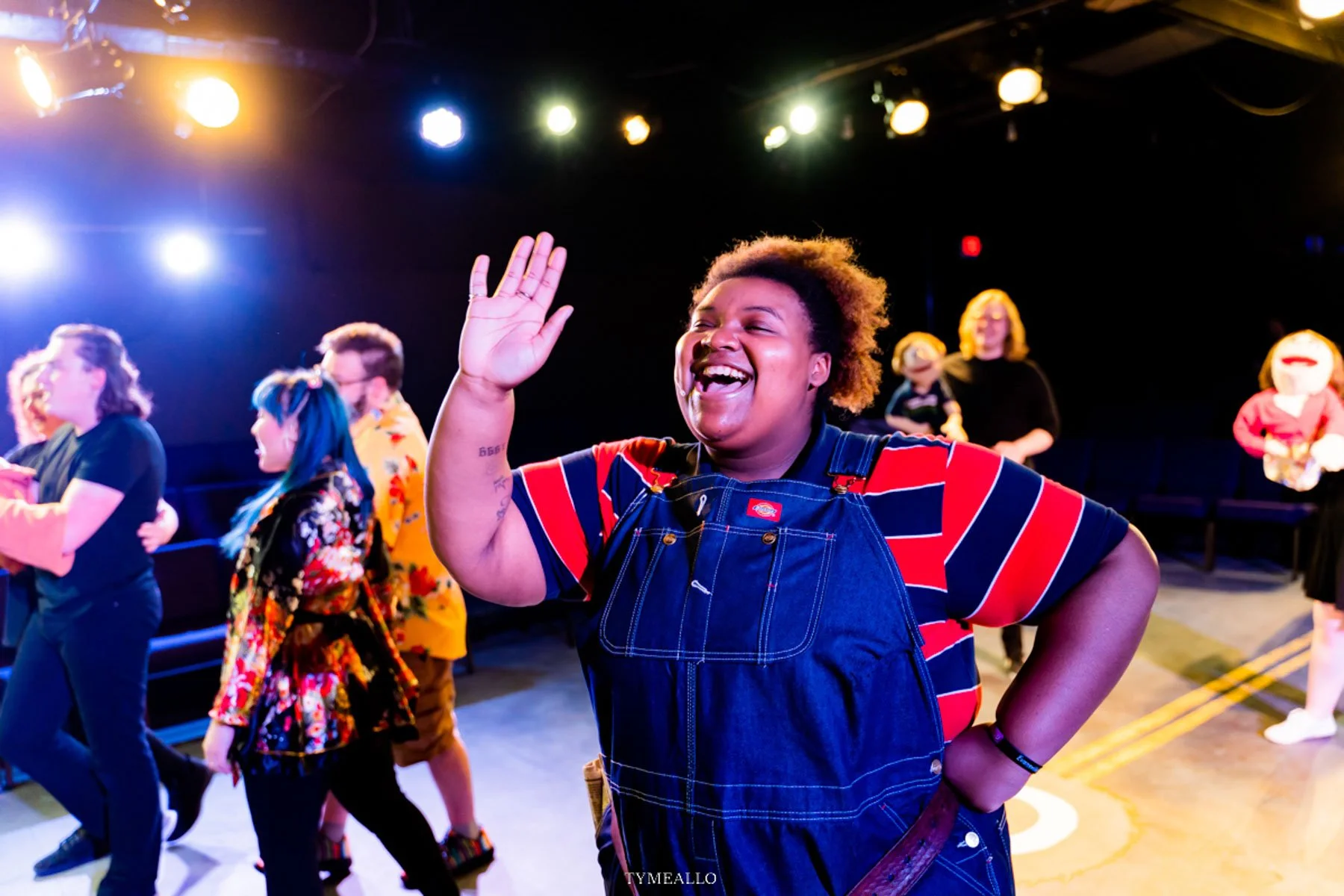 A woman with short curly hair, wearing a striped shirt and denim overalls, smiling and waving at a dance event. Behind her, other women are dancing and some are in costumes, set against a backdrop of stage lights.