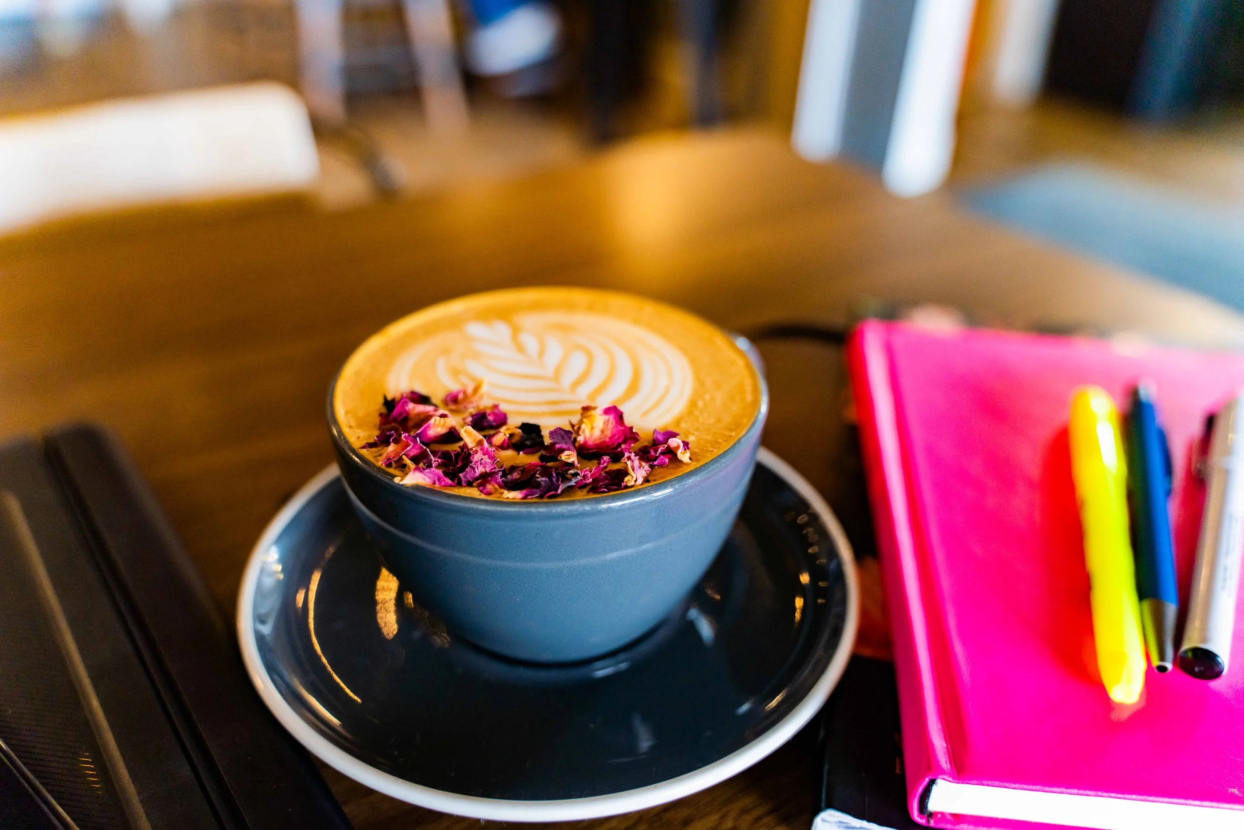 A cup of latte with latte art and pink flower petals on top, placed on a saucer on a wooden table, with colorful pens and a pink notebook nearby.