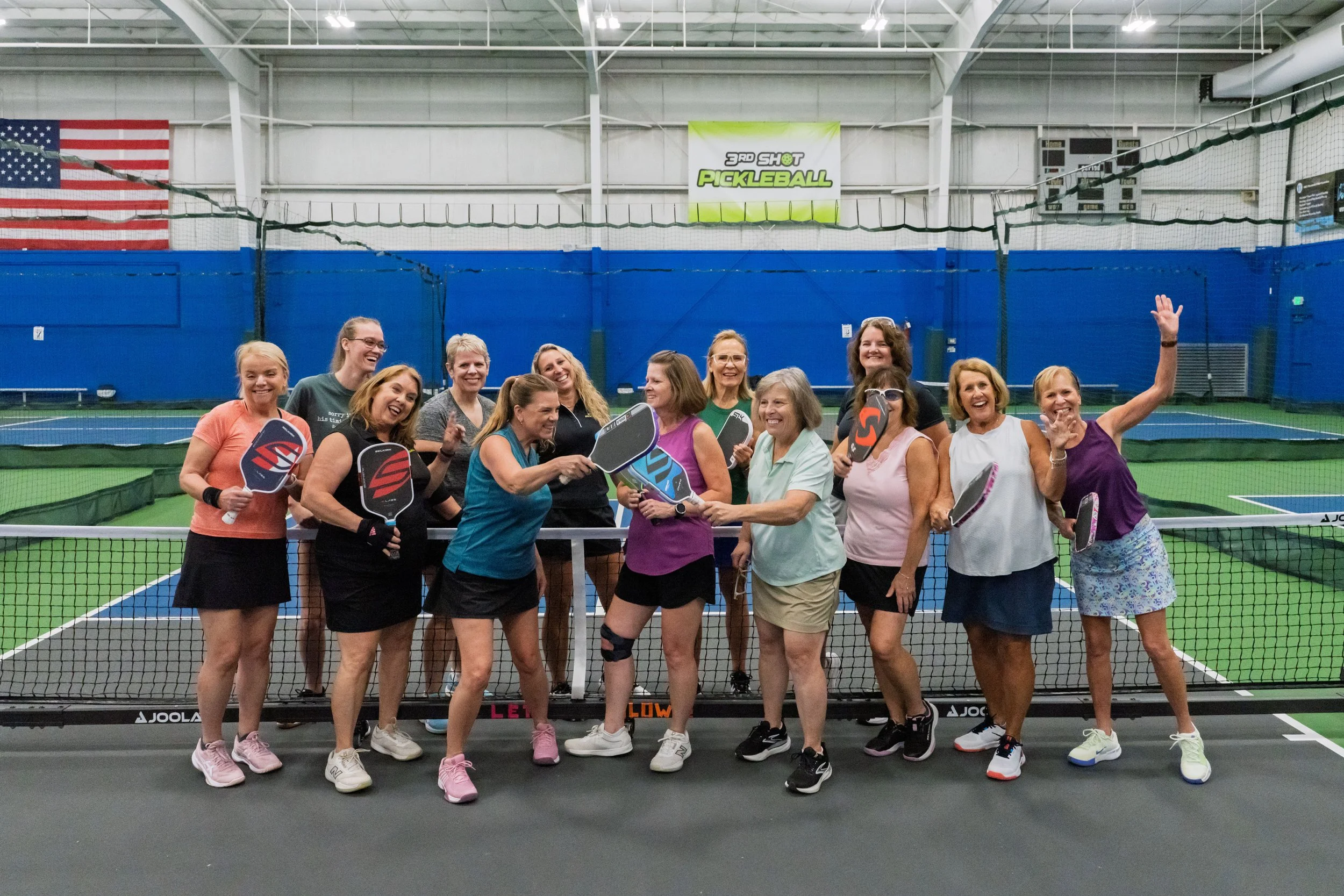 Group of women playing pickleball indoors, holding paddles, with a net and a blue wall in the background. An American flag is displayed on the left and a banner reading '3rd Shot Pickleball' hangs overhead.