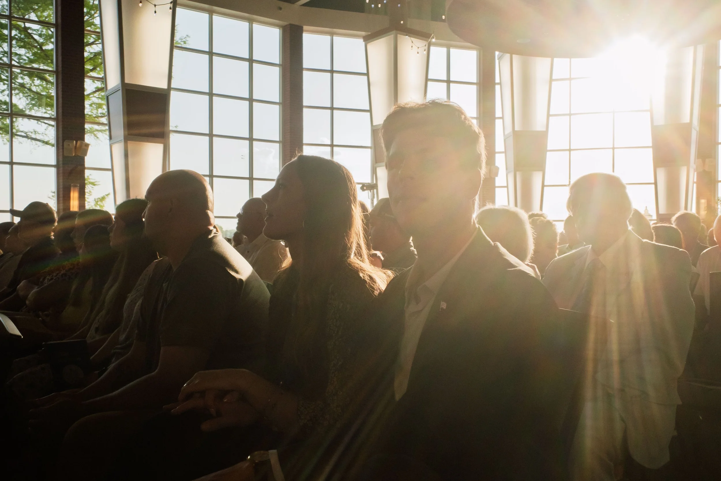 Silhouettes of people sitting and listening in a large room with tall windows and sunlight pouring in.