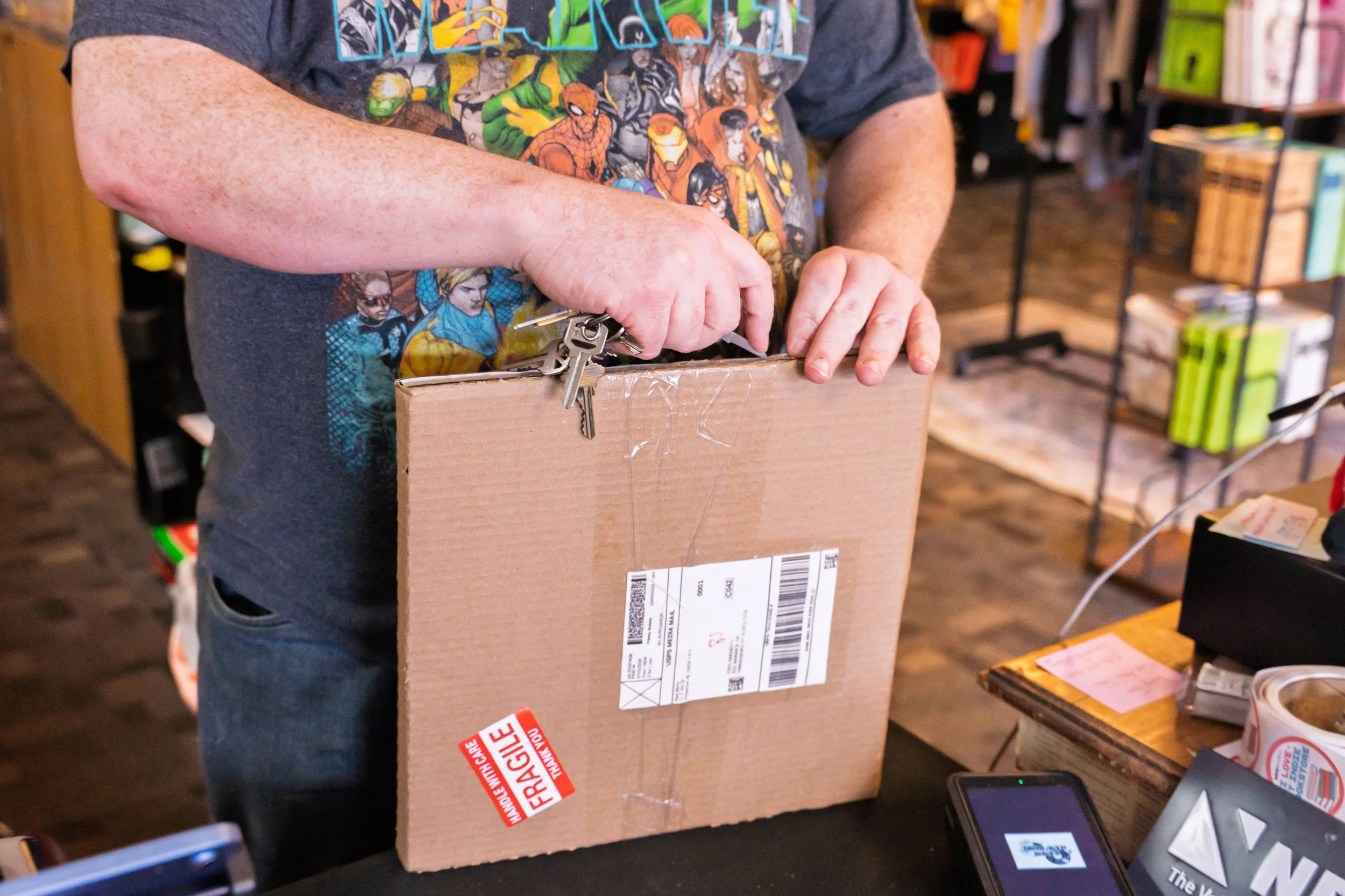 Person wearing a colorful superhero comic shirt is holding a cardboard box with shipping labels and a red "fragile" sticker on it.