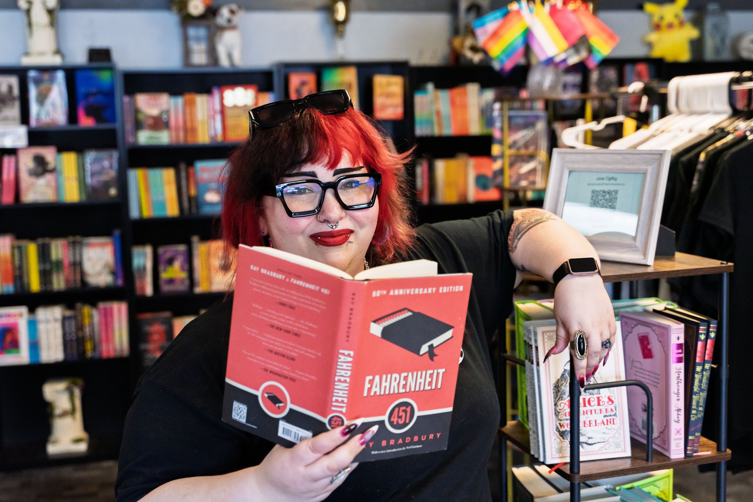 A woman with red and black hair and glasses reading a pink book titled 'Fahrenheit 451' in a bookstore.