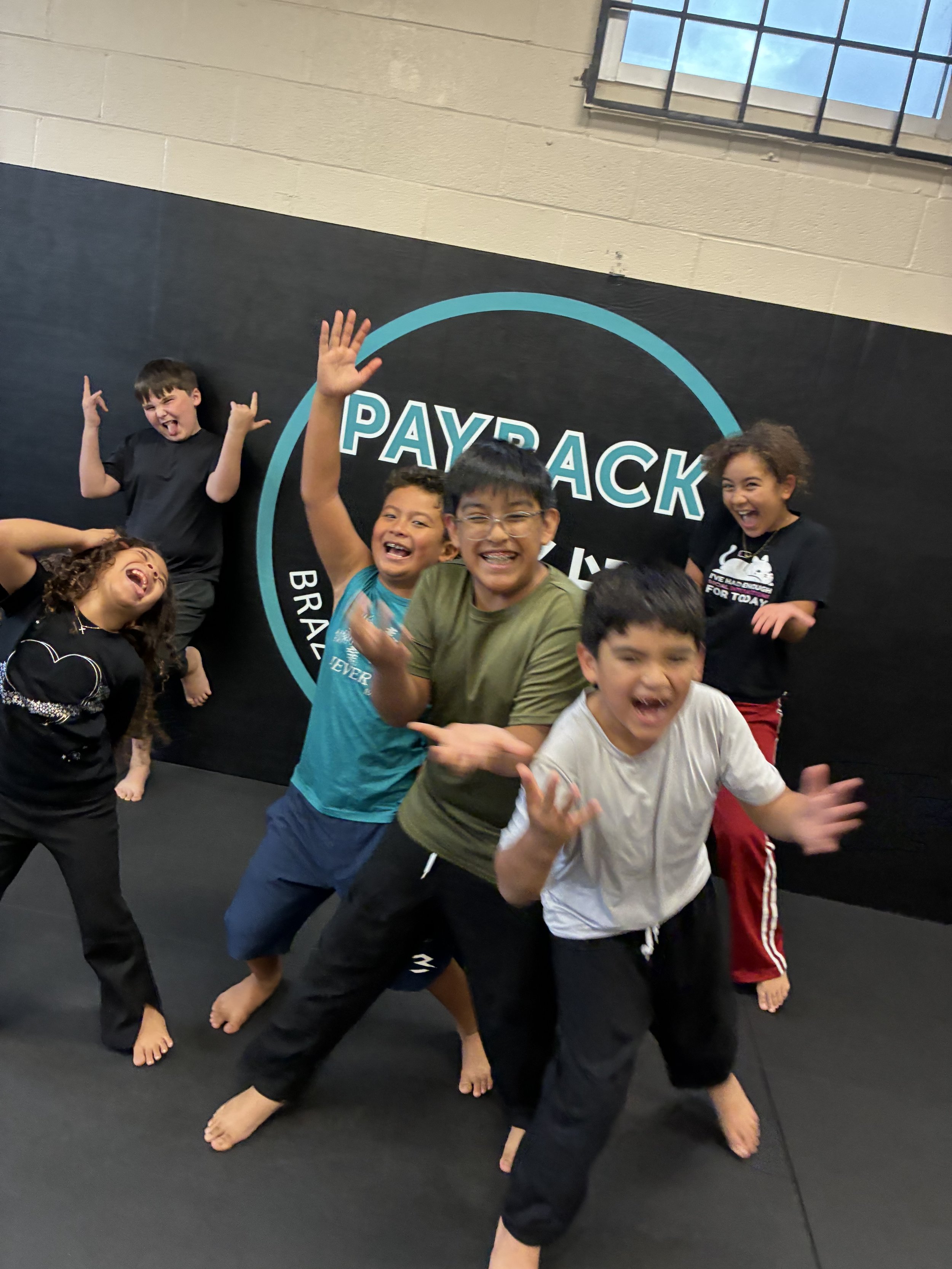 Group of children laughing and posing for a photo in a martial arts gym, with a large sign that says PAYBACK in the background.