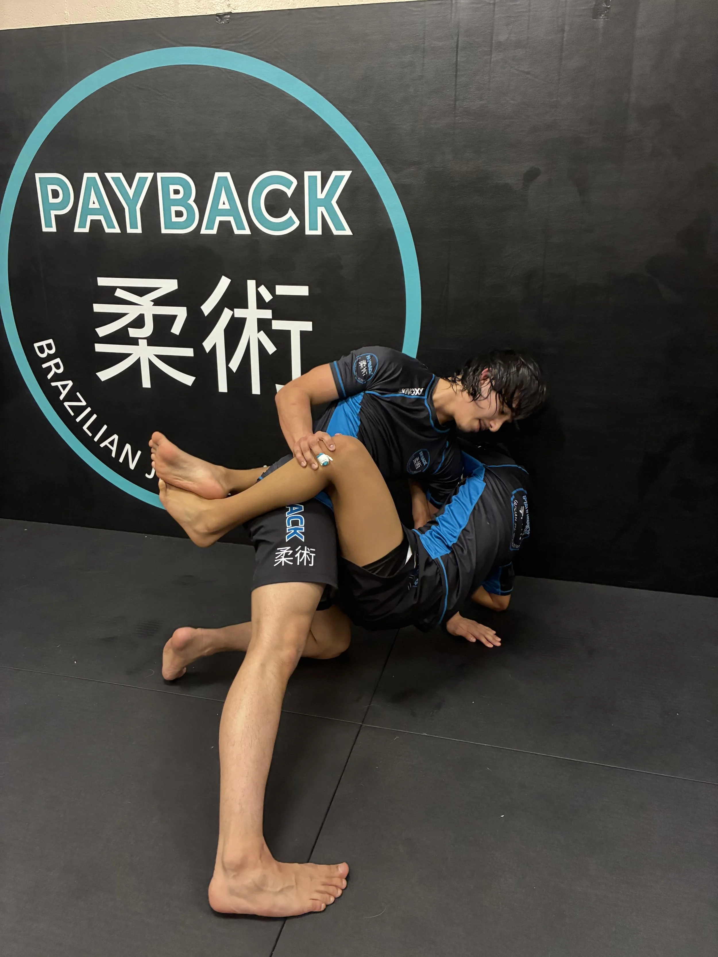 Two people practicing Brazilian Jiu-Jitsu grappling on a mat, with a black wall in the background displaying the words 'PAYBACK' and 'BRAZILIAN' with Japanese characters.