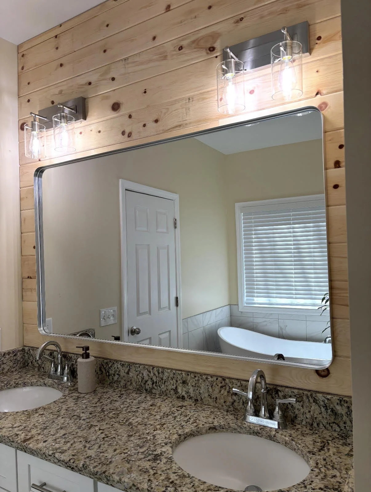 A bathroom mirror reflecting a window with blinds and a bathtub, mounted on a wood-paneled wall with light fixtures above.