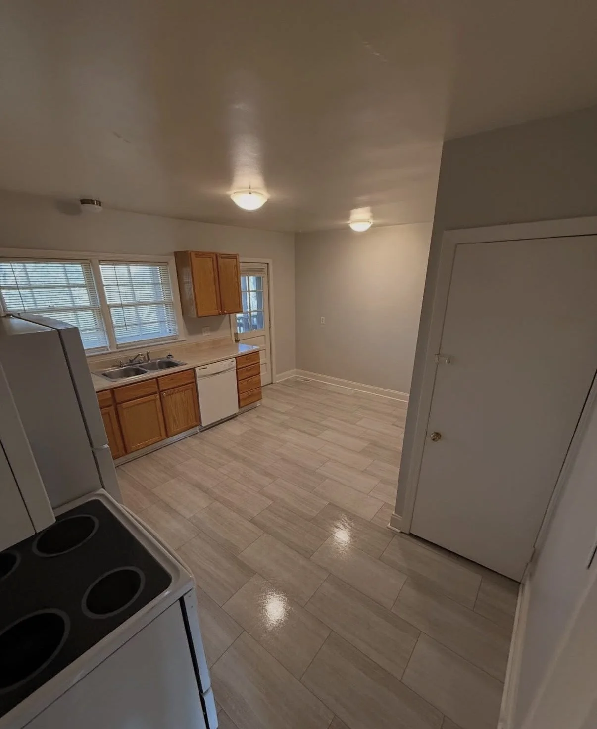 Empty kitchen with wooden cabinets, white dishwasher, stove, and windowed door. Light-colored floor tiles, gray walls, ceiling lights, and a small white door.